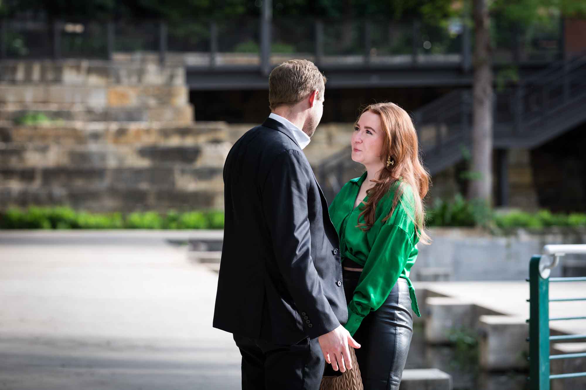 Man wearing black suit standing in front of woman wearing green blouse on sidewalk in front of Emma Hotel beside Riverwalk during an Emma Hotel surprise proposal