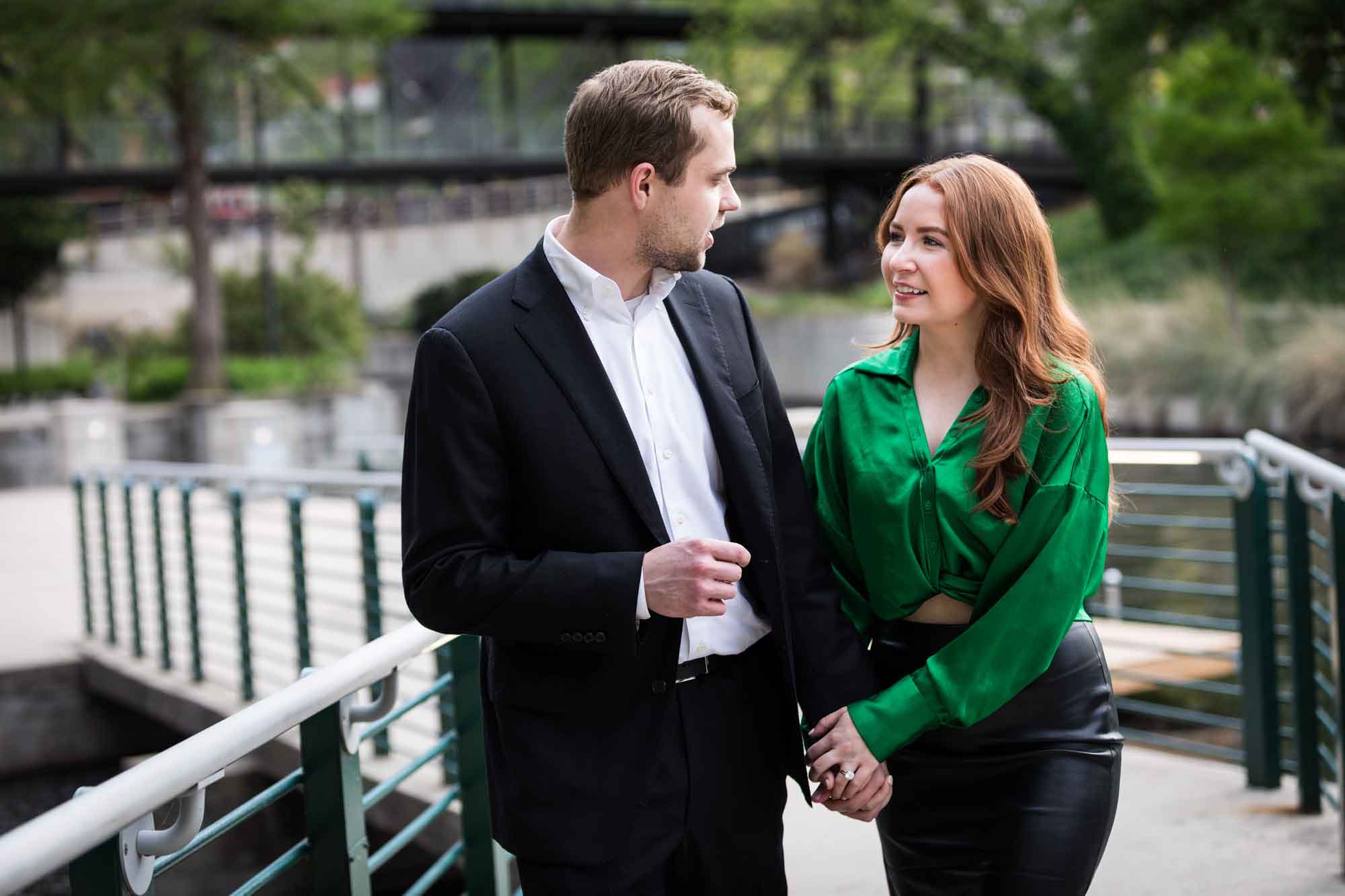 Man wearing black suit walking with woman wearing green blouse along railing beside Riverwalk during an Emma Hotel surprise proposal