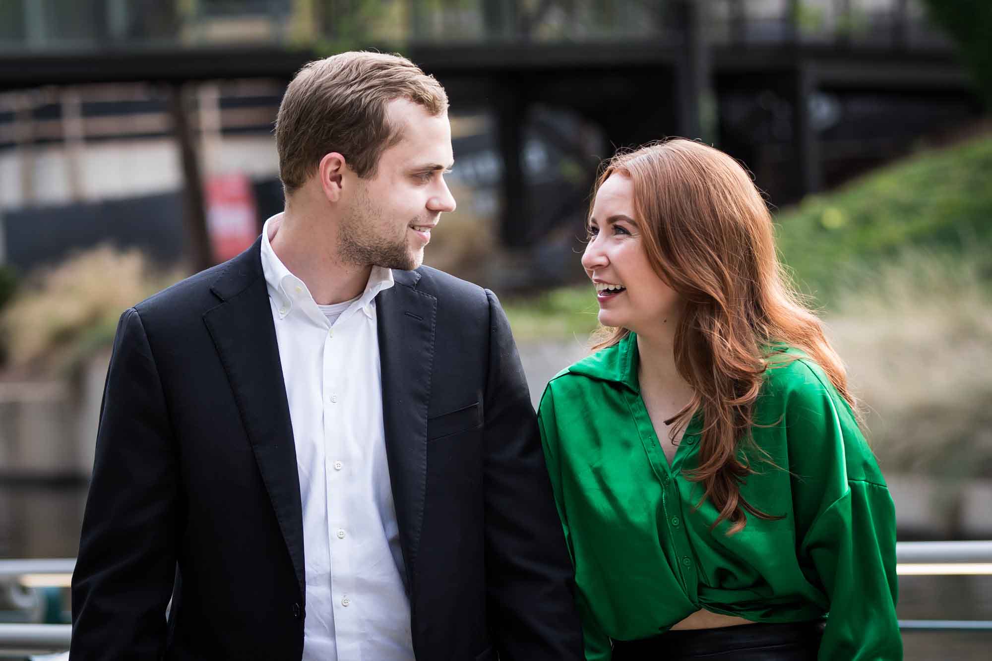 Man wearing black suit walking with woman wearing green blouse beside Riverwalk during an Emma Hotel surprise proposal