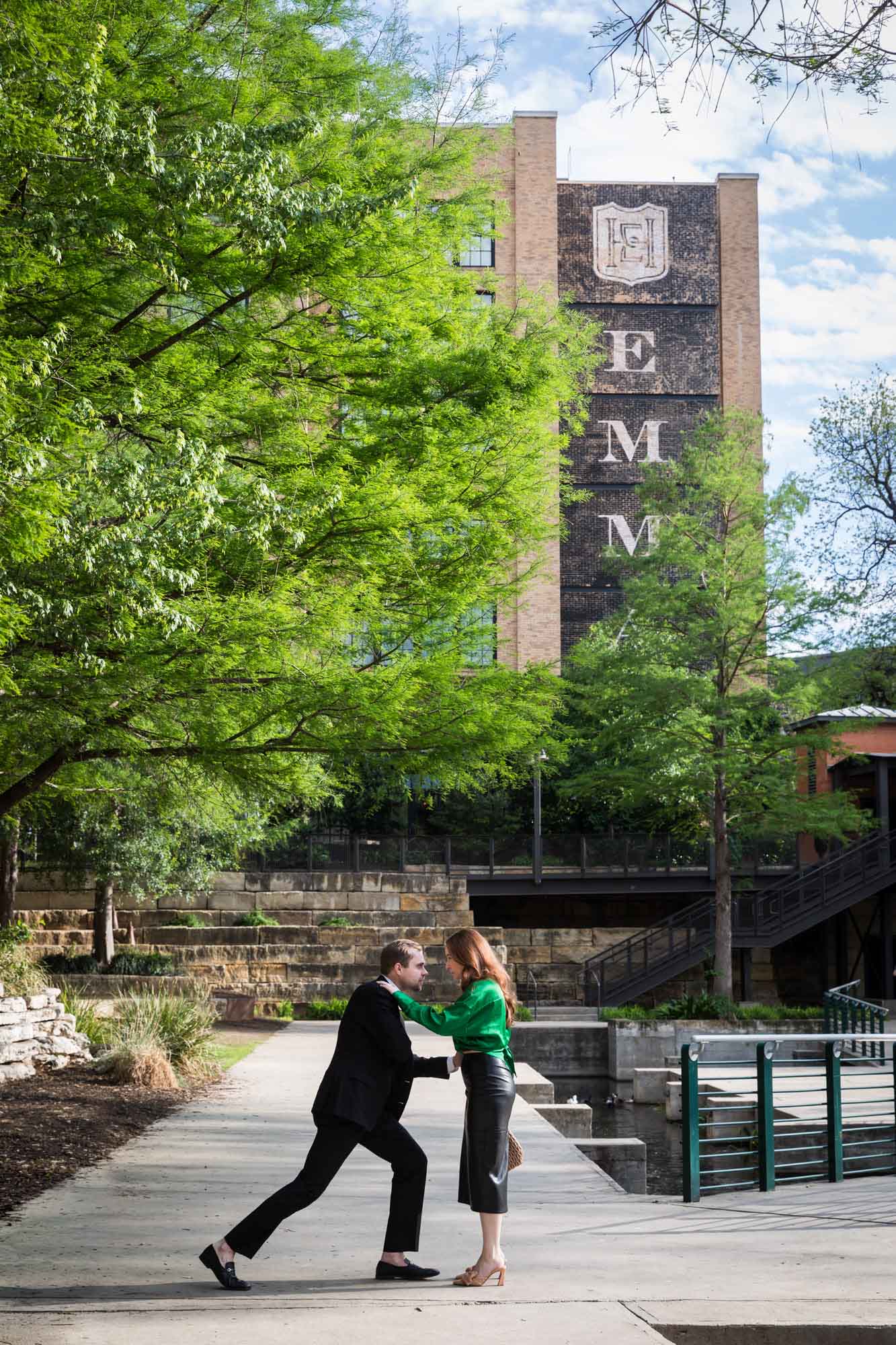 Man wearing black suit getting down on one knee in front of woman wearing green blouse on sidewalk in front of Emma Hotel beside Riverwalk during an Emma Hotel surprise proposal