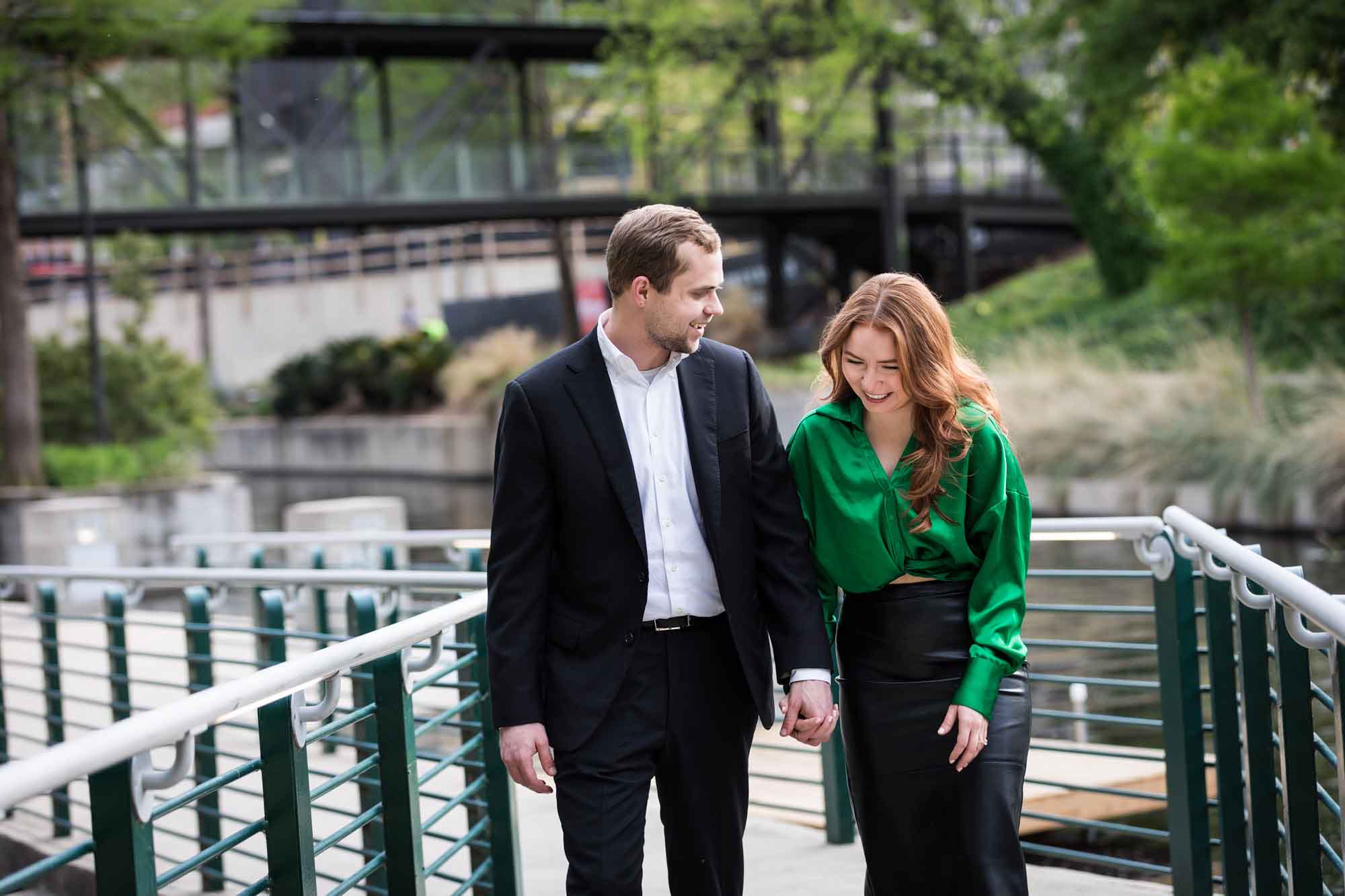 Man wearing black suit walking with woman wearing green blouse along railing beside Riverwalk during an Emma Hotel surprise proposal
