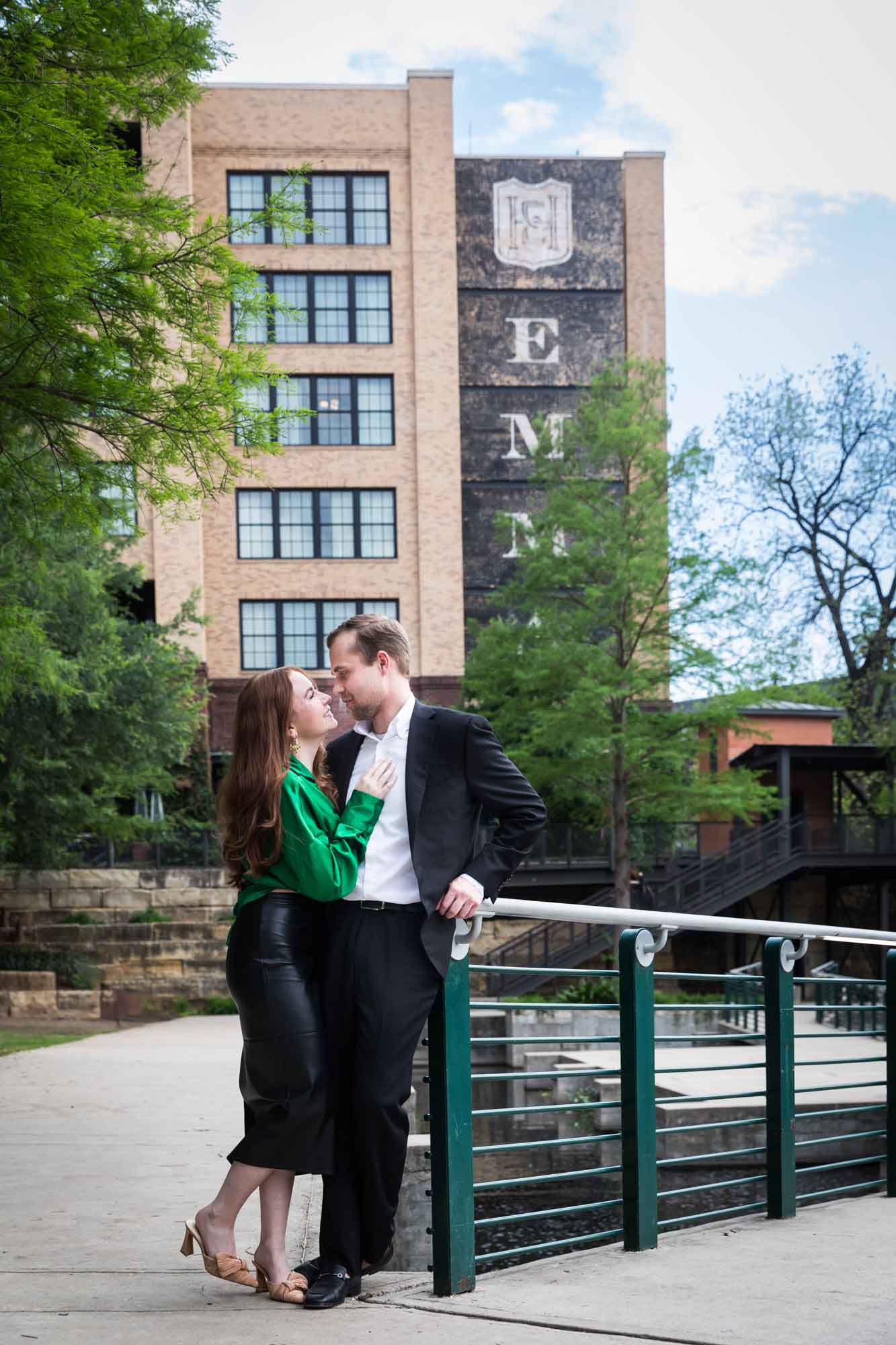 Man wearing black suit walking with woman wearing green blouse standing in front of railing beside Riverwalk during an Emma Hotel surprise proposal