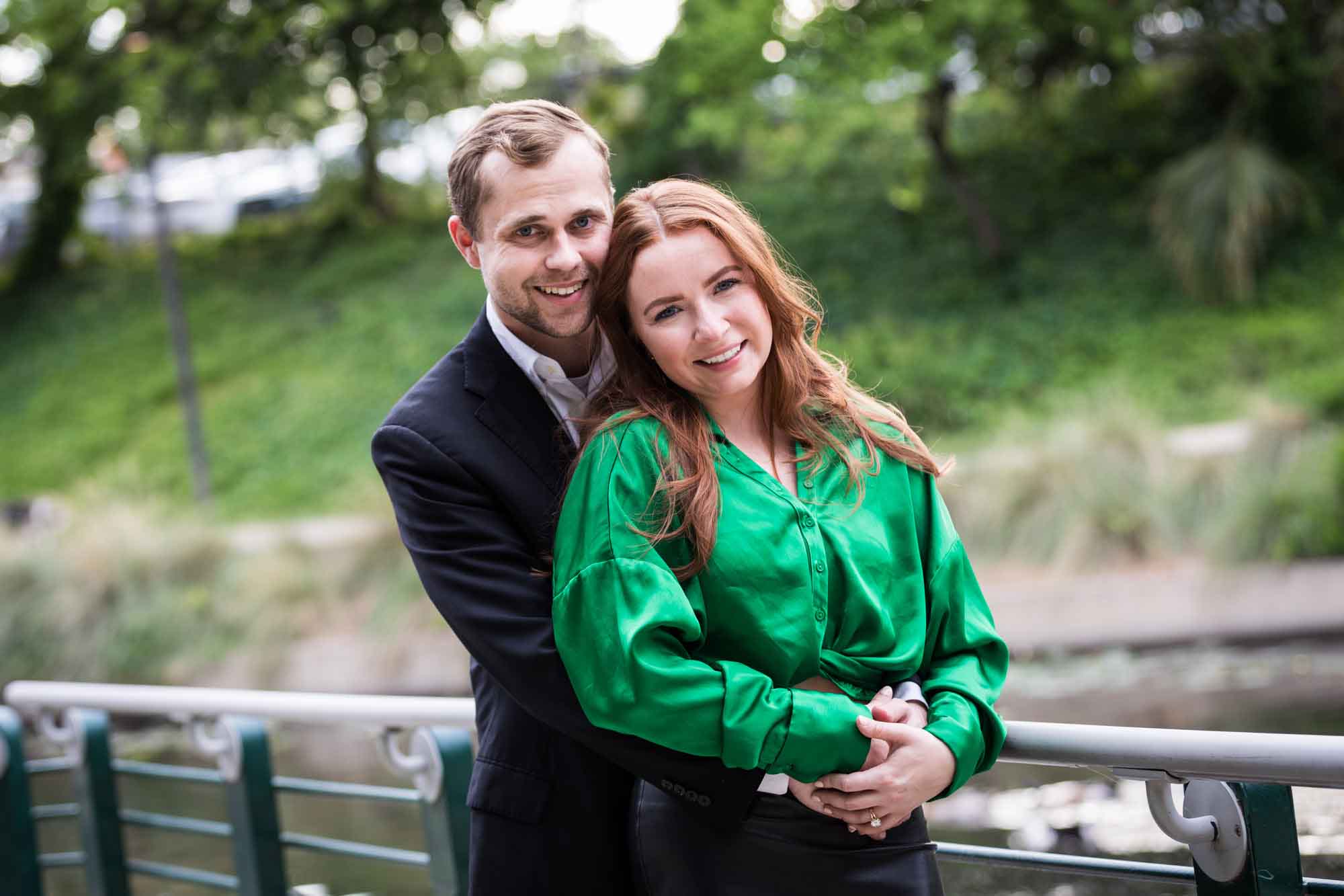 Man wearing black suit standing with woman wearing green blouse along railing beside Riverwalk during an Emma Hotel surprise proposal