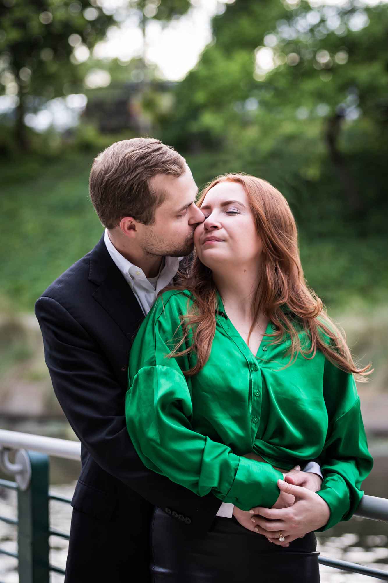 Man wearing black suit kissing woman wearing green blouse along railing beside Riverwalk during an Emma Hotel surprise proposal