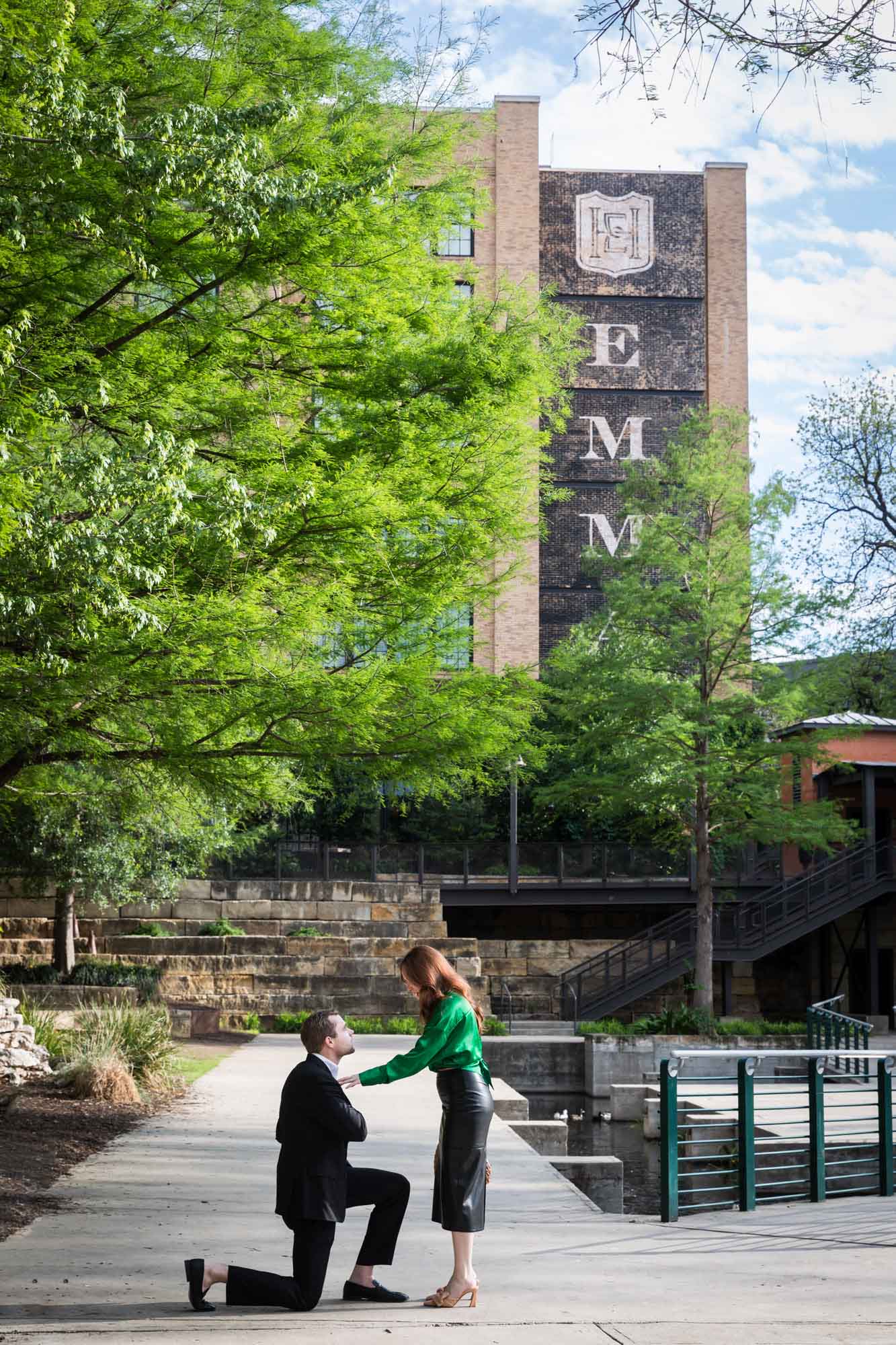 Man wearing black suit proposing while down on one knee in front of woman wearing green blouse on sidewalk in front of Emma Hotel beside Riverwalk during an Emma Hotel surprise proposal