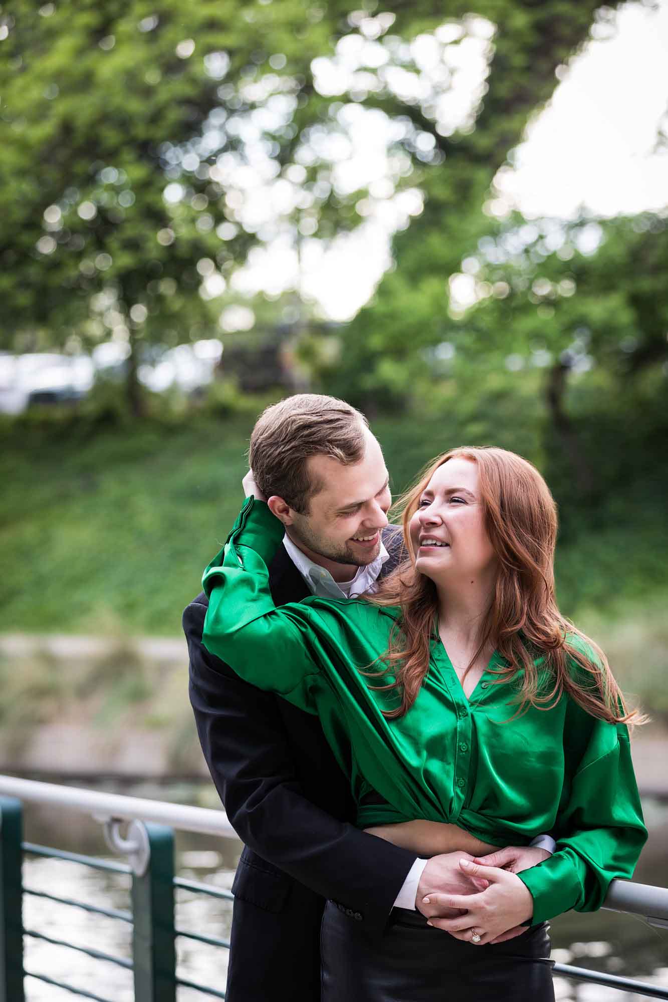 Man wearing black suit standing with woman wearing green blouse along railing beside Riverwalk during an Emma Hotel surprise proposal