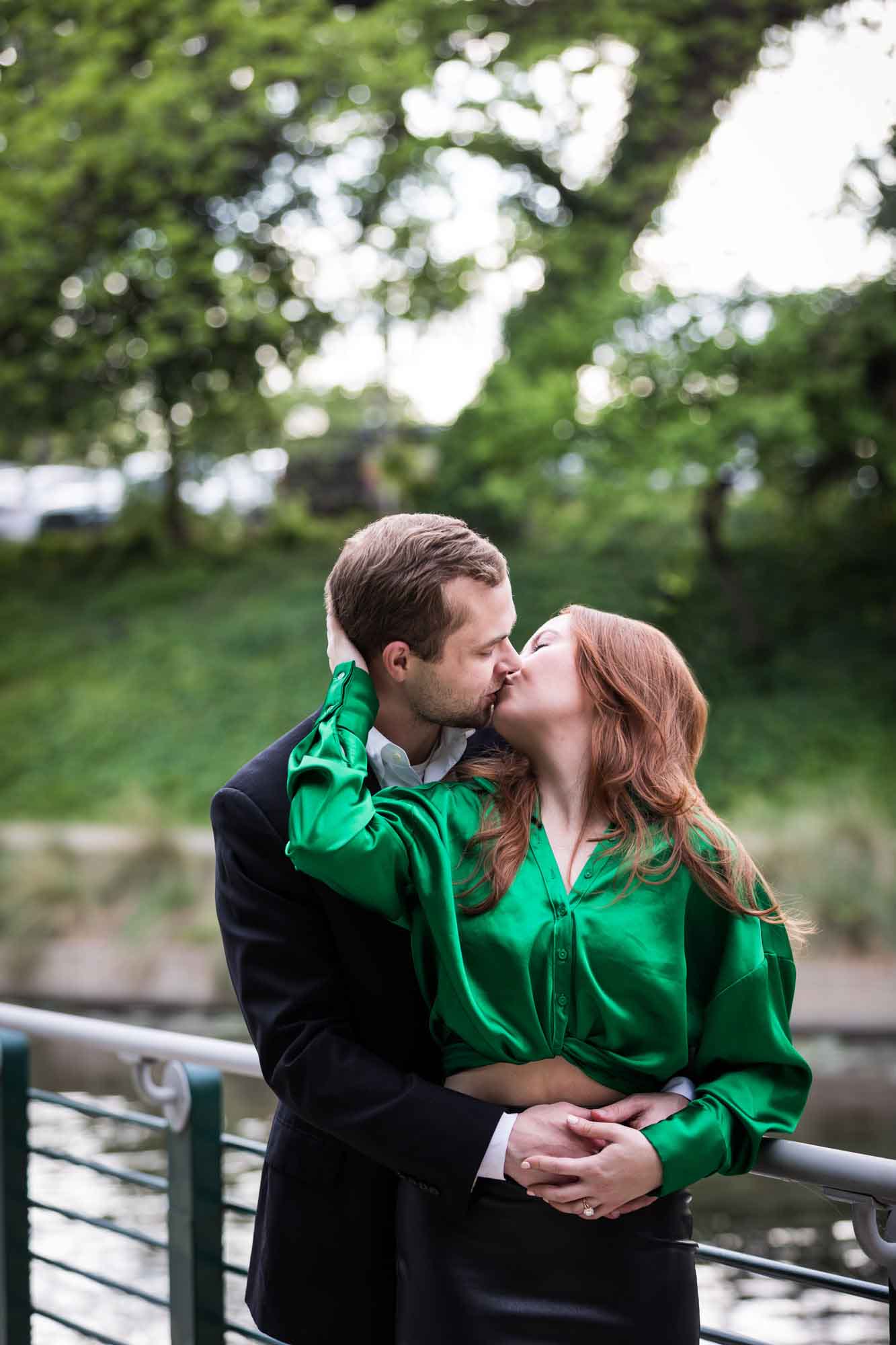 Man wearing black suit kissing woman wearing green blouse along railing beside Riverwalk during an Emma Hotel surprise proposal