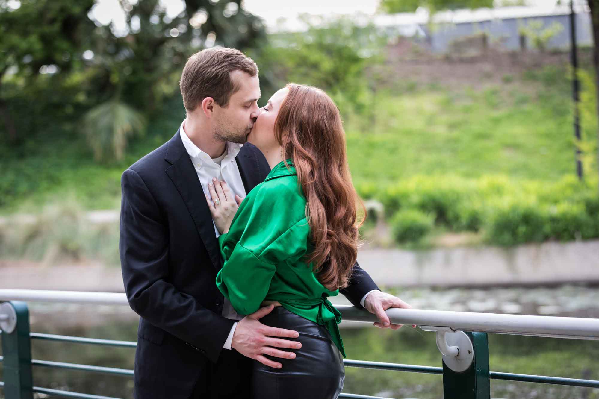 Man wearing black suit kissing woman wearing green blouse along railing beside Riverwalk during an Emma Hotel surprise proposal