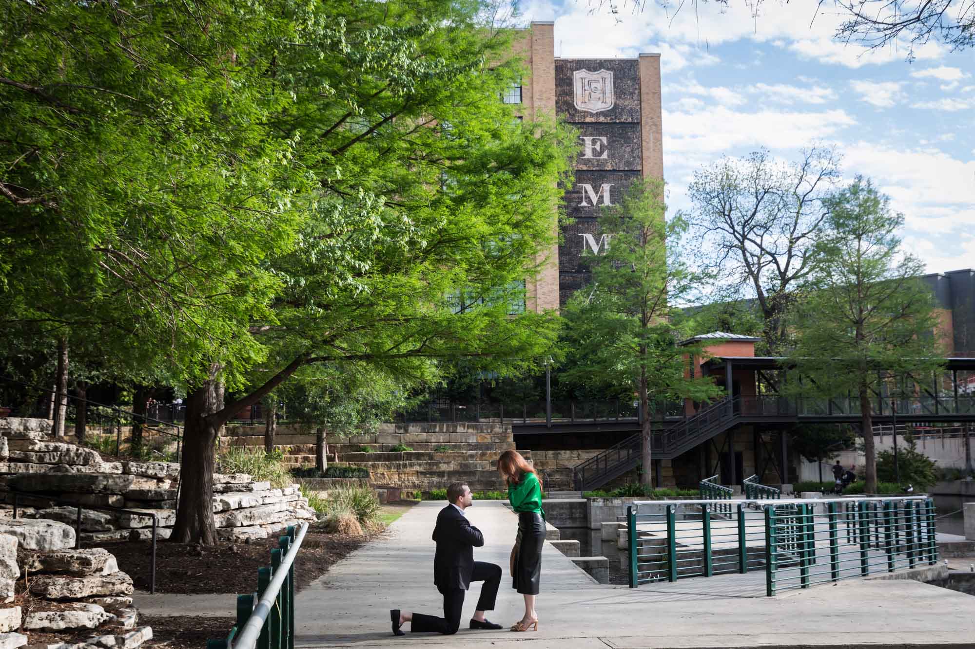 Man wearing black suit proposing while down on one knee in front of woman wearing green blouse on sidewalk in front of Emma Hotel beside Riverwalk during an Emma Hotel surprise proposal
