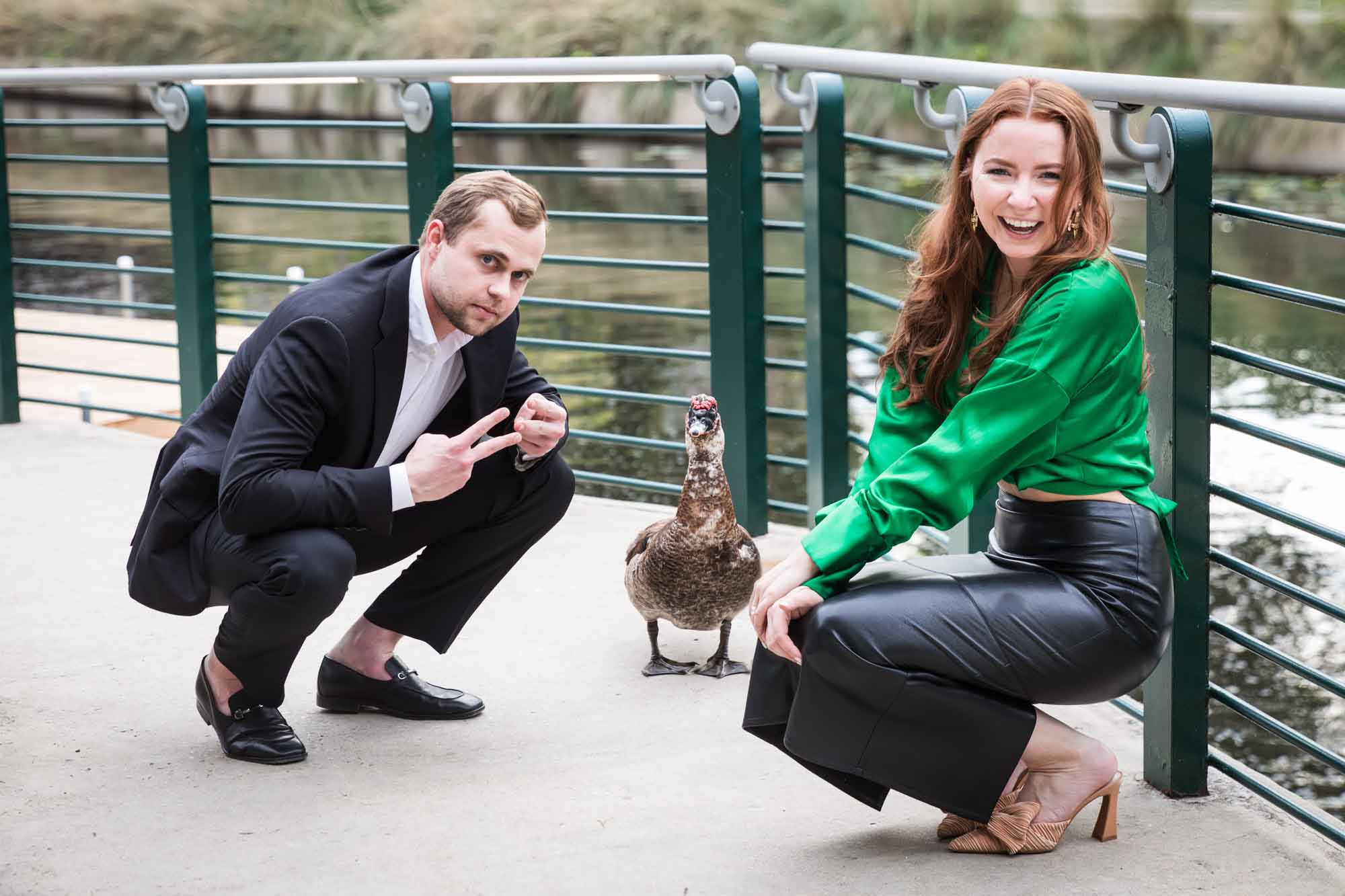 Man wearing black suit squatting with woman wearing green blouse along railing beside Riverwalk with duck during an Emma Hotel proposal
