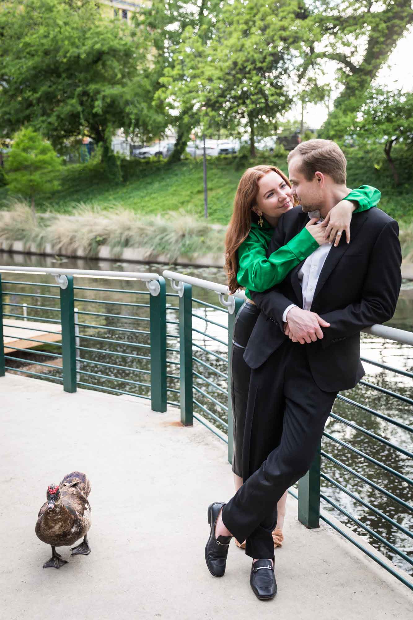 Man wearing black suit being hugged by woman wearing green blouse along railing beside Riverwalk with duck walking past during an Emma Hotel proposal