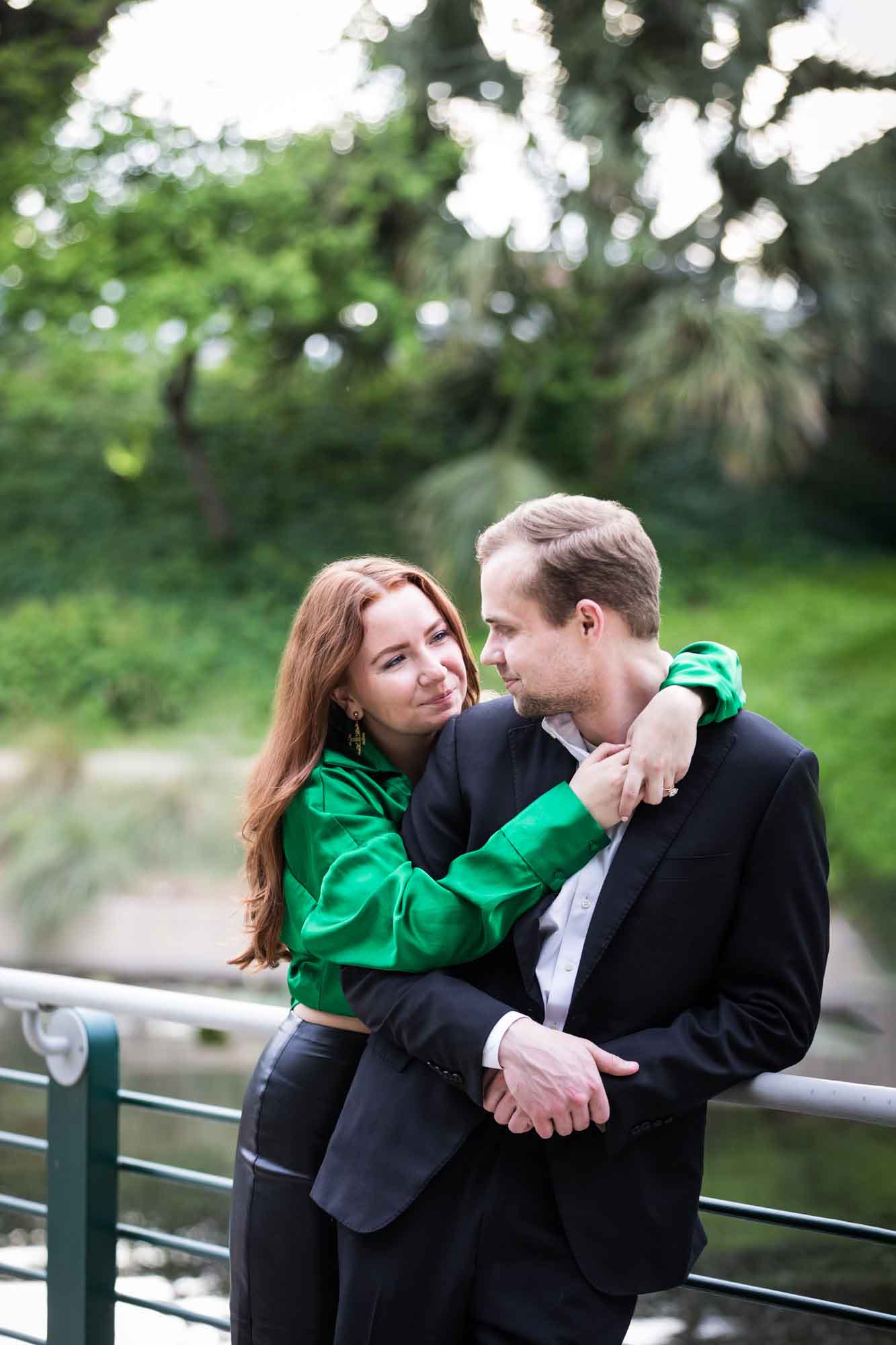 Man wearing black suit being hugged by woman wearing green blouse along railing beside Riverwalk during an Emma Hotel proposal