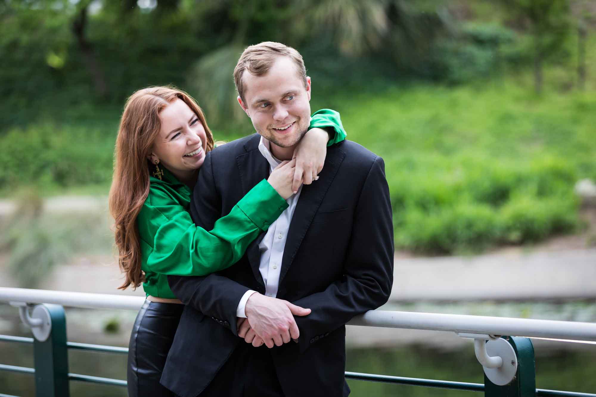 Man wearing black suit being hugged by woman wearing green blouse along railing beside Riverwalk during an Emma Hotel proposal