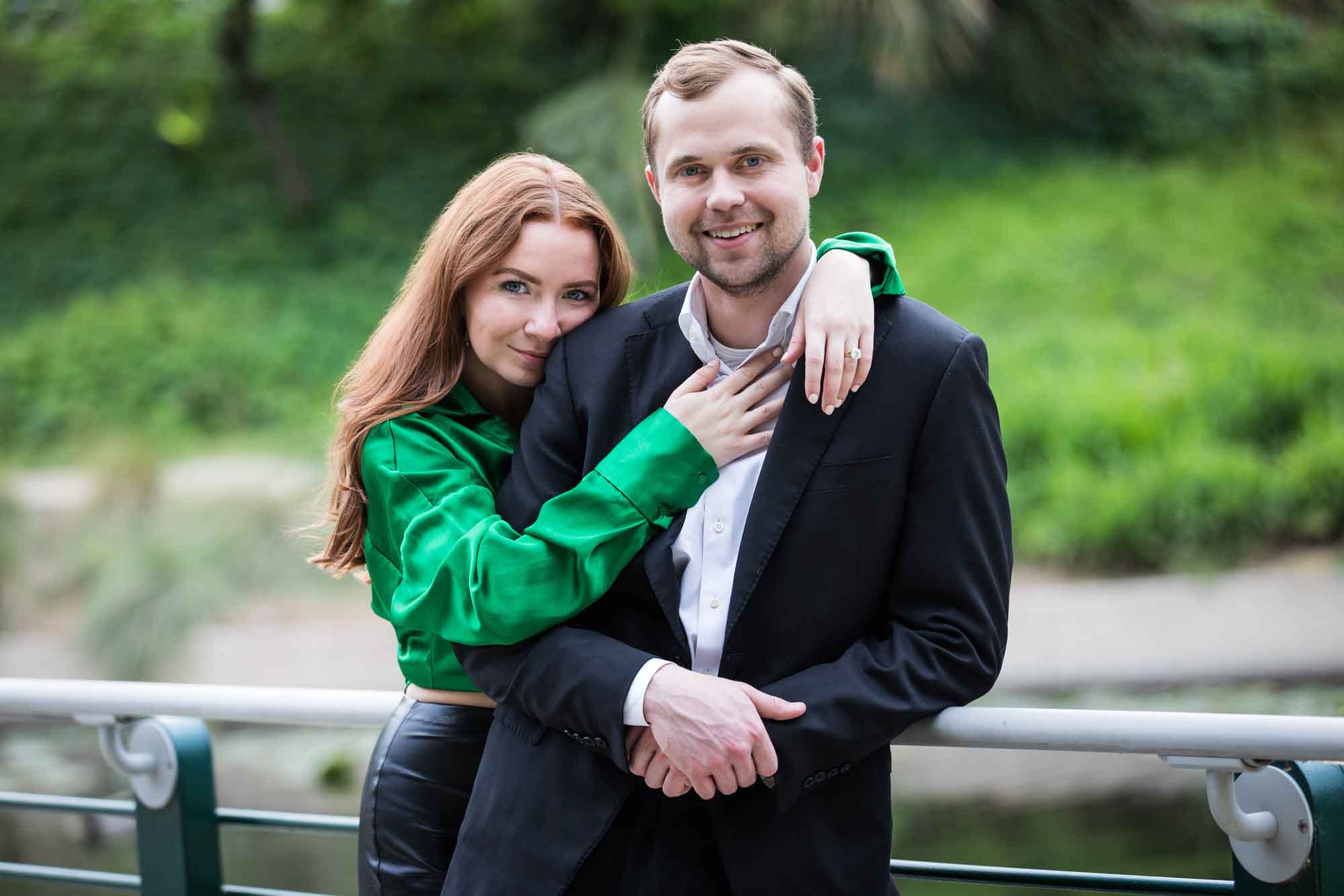 Man wearing black suit being hugged by woman wearing green blouse along railing beside Riverwalk during an Emma Hotel proposal