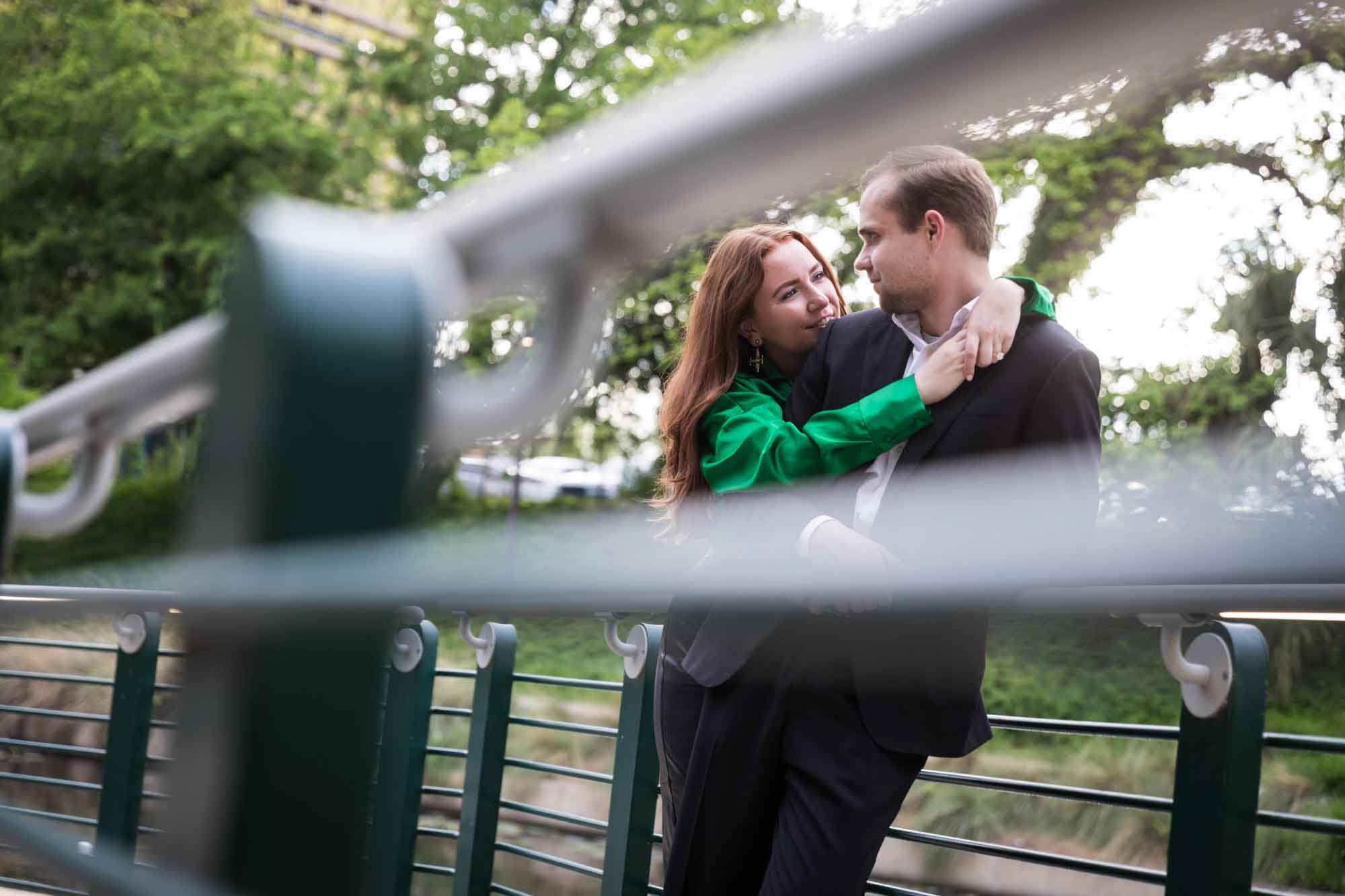 Man wearing black suit being hugged by woman wearing green blouse along railing beside Riverwalk during an Emma Hotel proposal