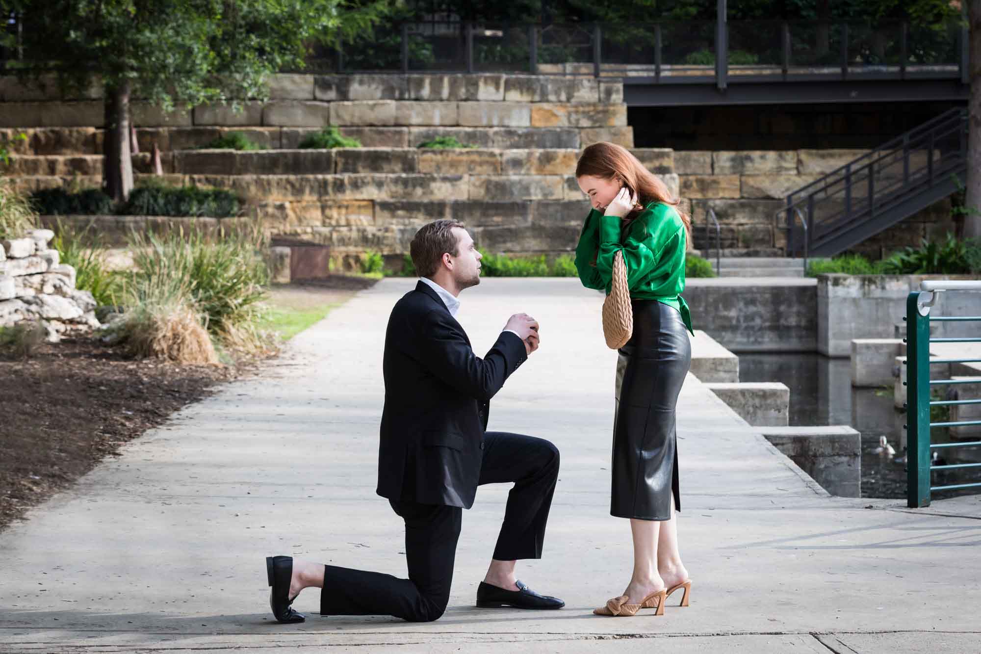 Man wearing black suit proposing while down on one knee in front of woman wearing green blouse on sidewalk in front of Emma Hotel beside Riverwalk during an Emma Hotel surprise proposal