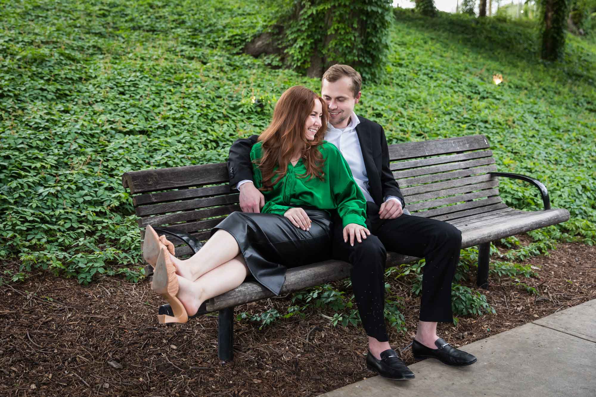 Man wearing black suit sitting on wooden bench with woman wearing green blouse in front of vine-covered hill during an Emma Hotel proposal
