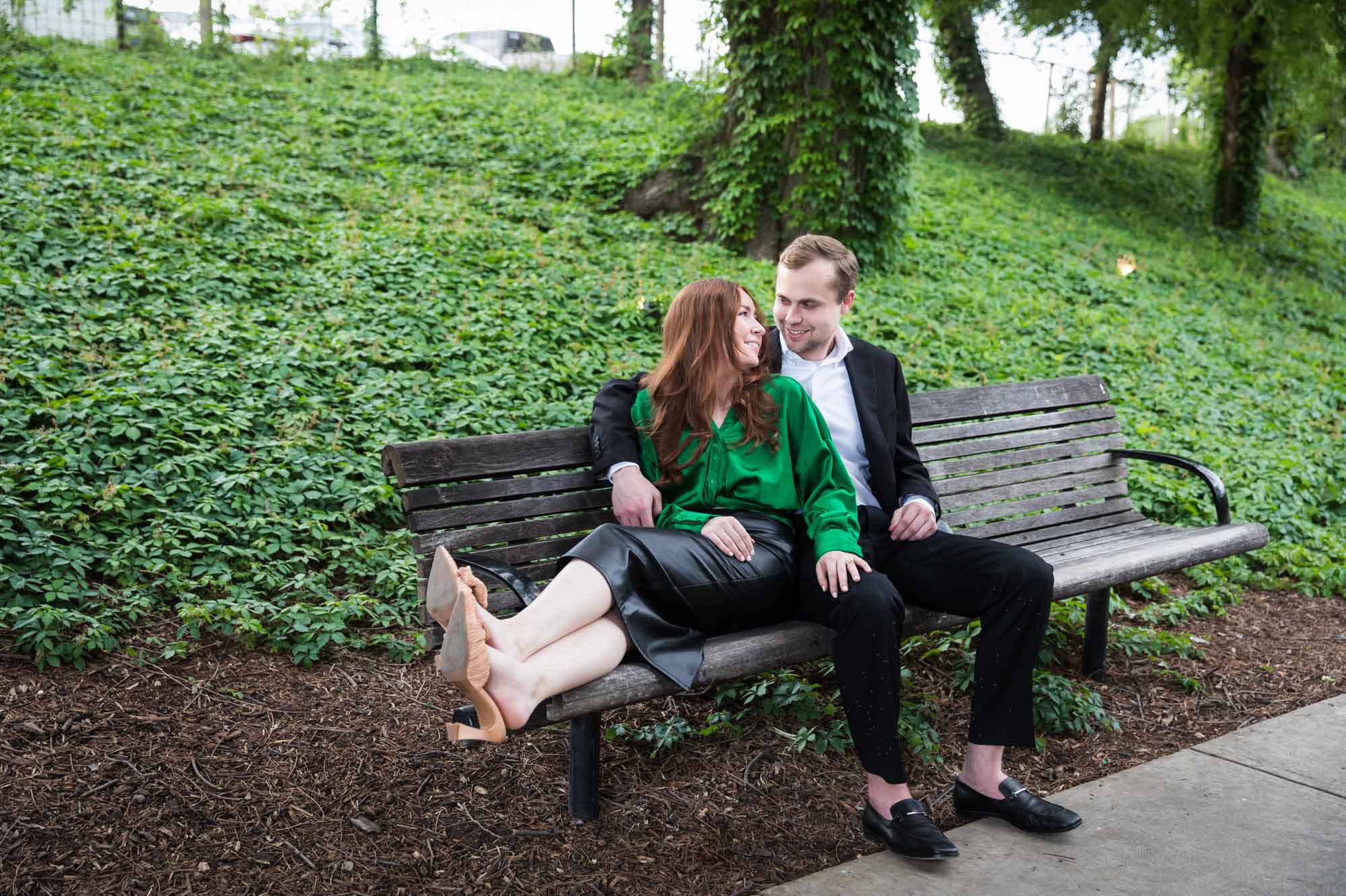 Man wearing black suit sitting on wooden bench with woman wearing green blouse in front of vine-covered hill during an Emma Hotel proposal