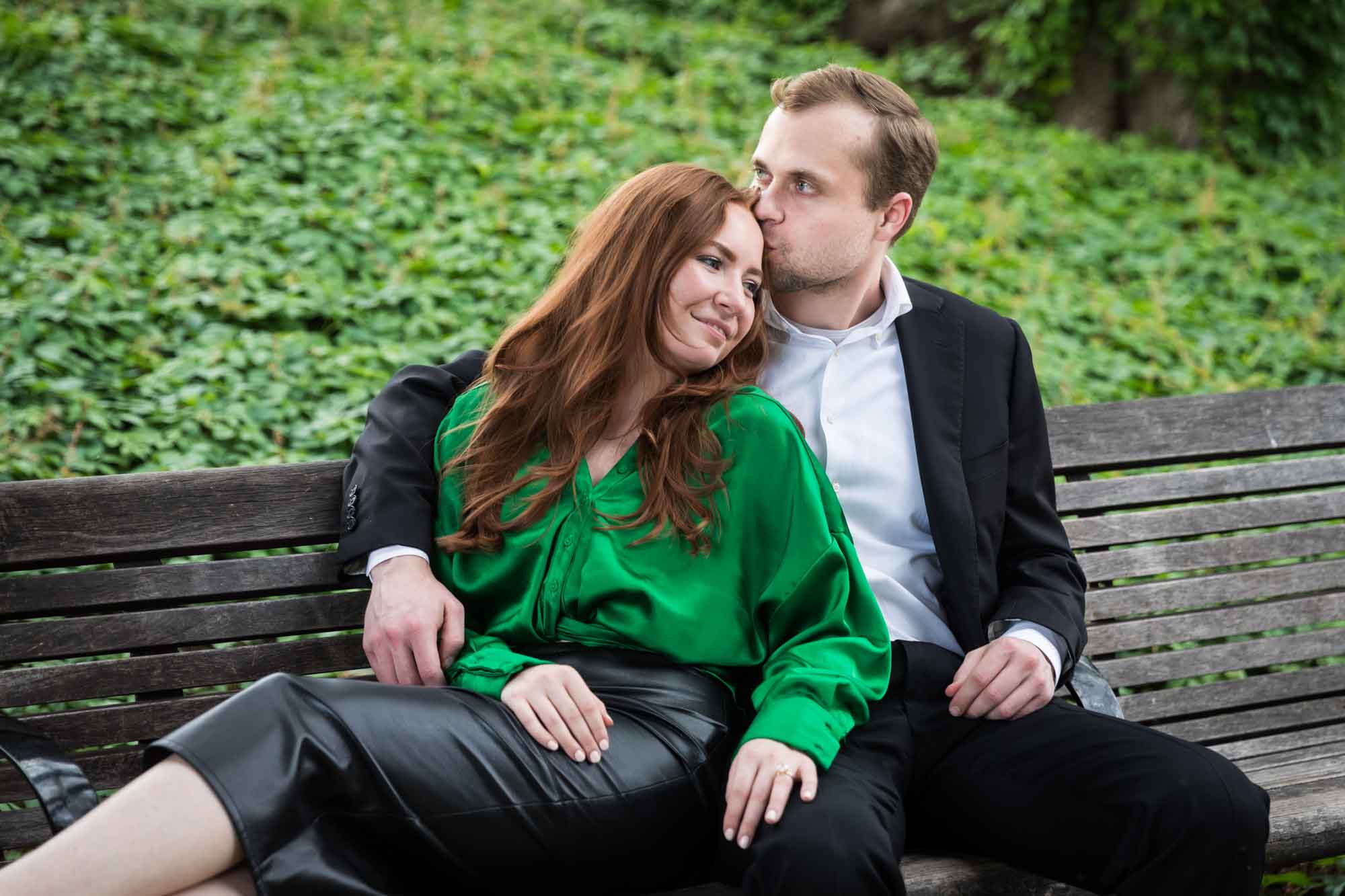 Man wearing black suit sitting on wooden bench kissing woman wearing green blouse in front of vine-covered hill during an Emma Hotel proposal