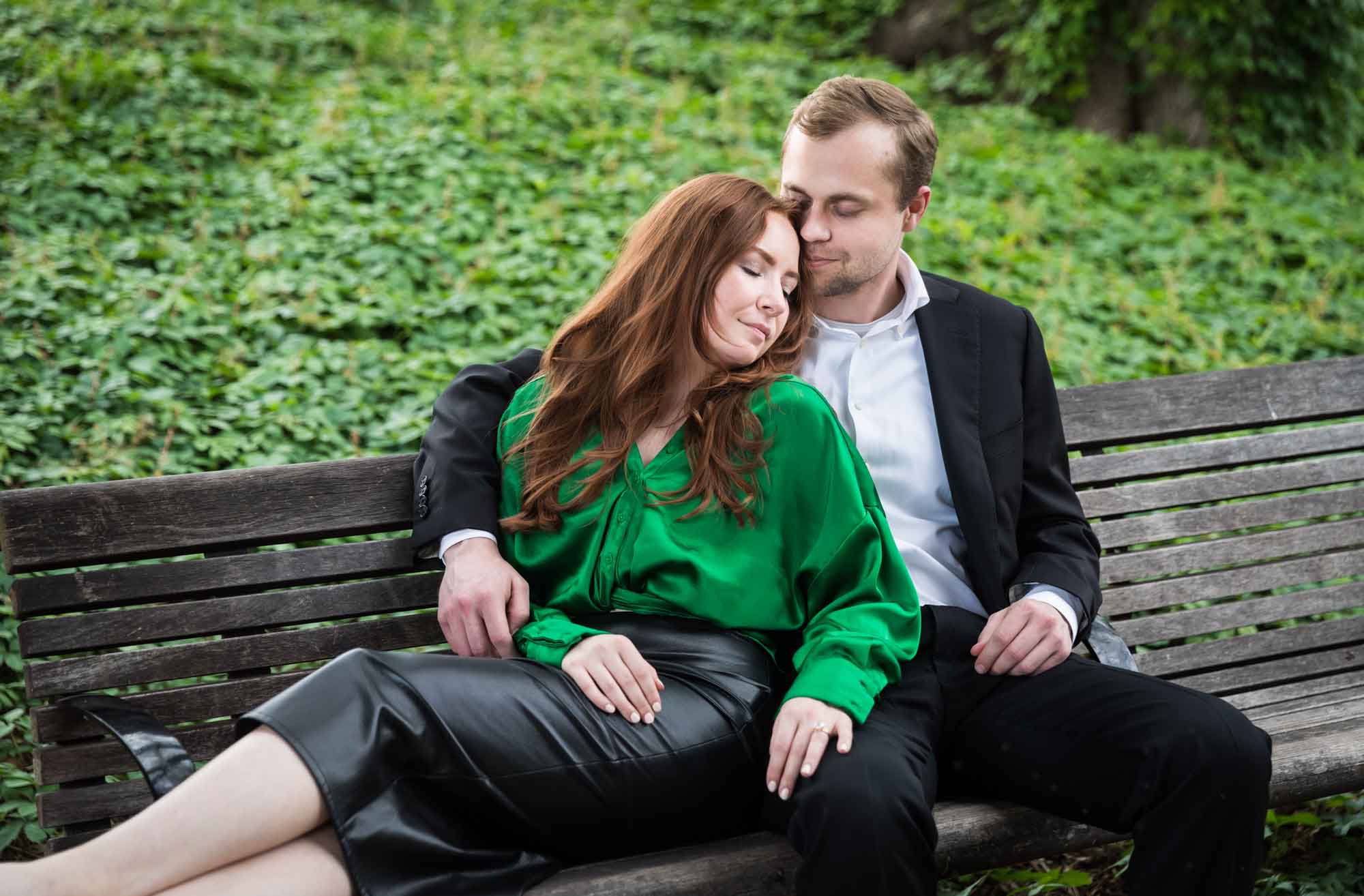 Man wearing black suit sitting on wooden bench with woman wearing green blouse in front of vine-covered hill during an Emma Hotel proposal