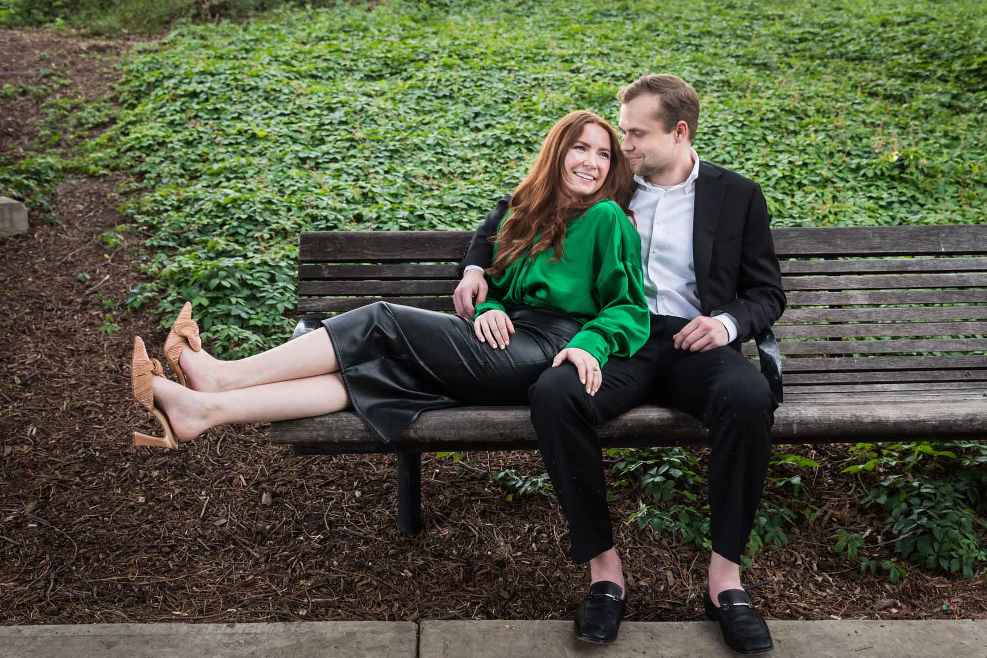 Man wearing black suit sitting on wooden bench with woman wearing green blouse in front of vine-covered hill during an Emma Hotel proposal