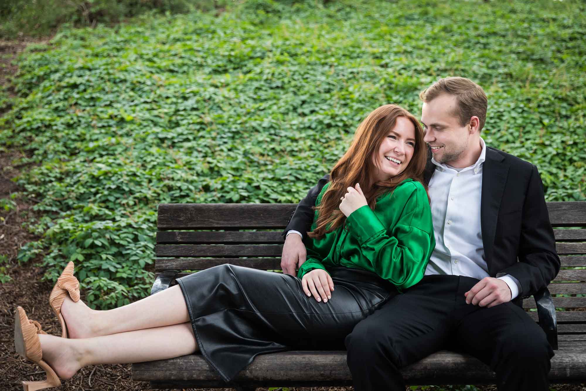 Man wearing black suit sitting on wooden bench with woman wearing green blouse in front of vine-covered hill during an Emma Hotel proposal