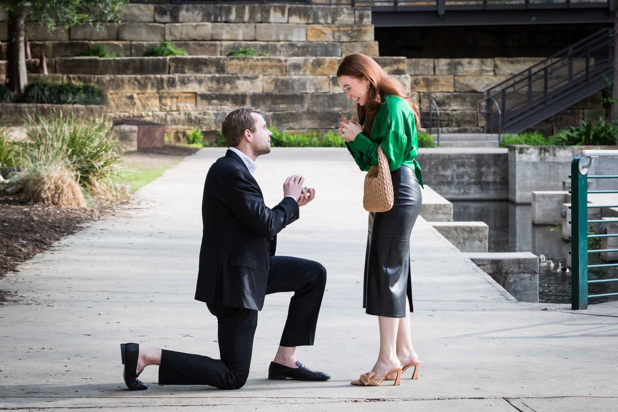 Man wearing black suit proposing while down on one knee in front of woman wearing green blouse on sidewalk in front of Emma Hotel beside Riverwalk during an Emma Hotel surprise proposal