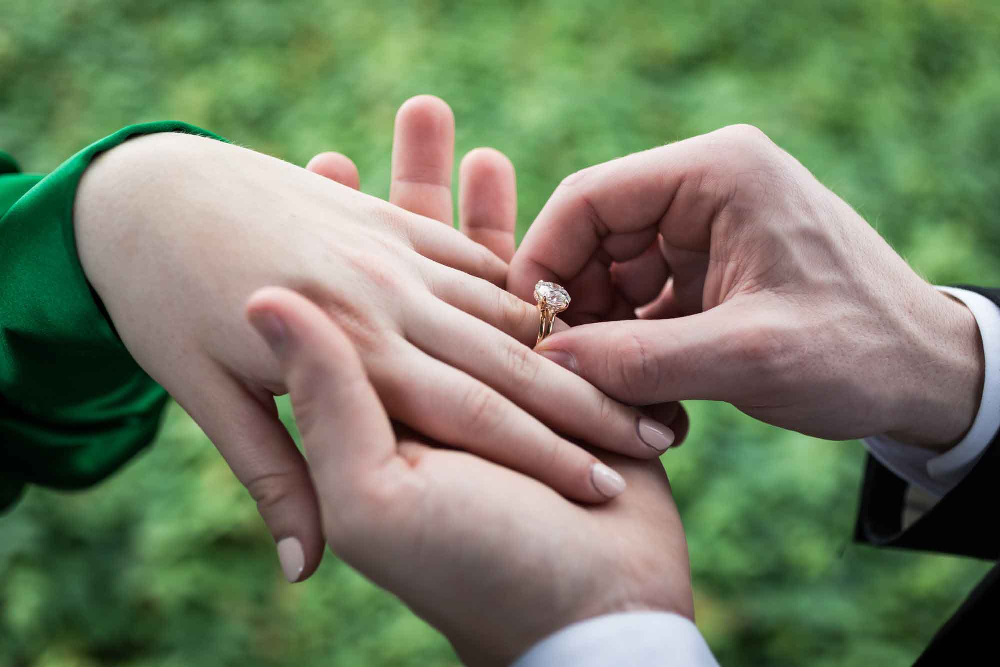 Close up of man's hand putting engagement ring on woman's hand with green background