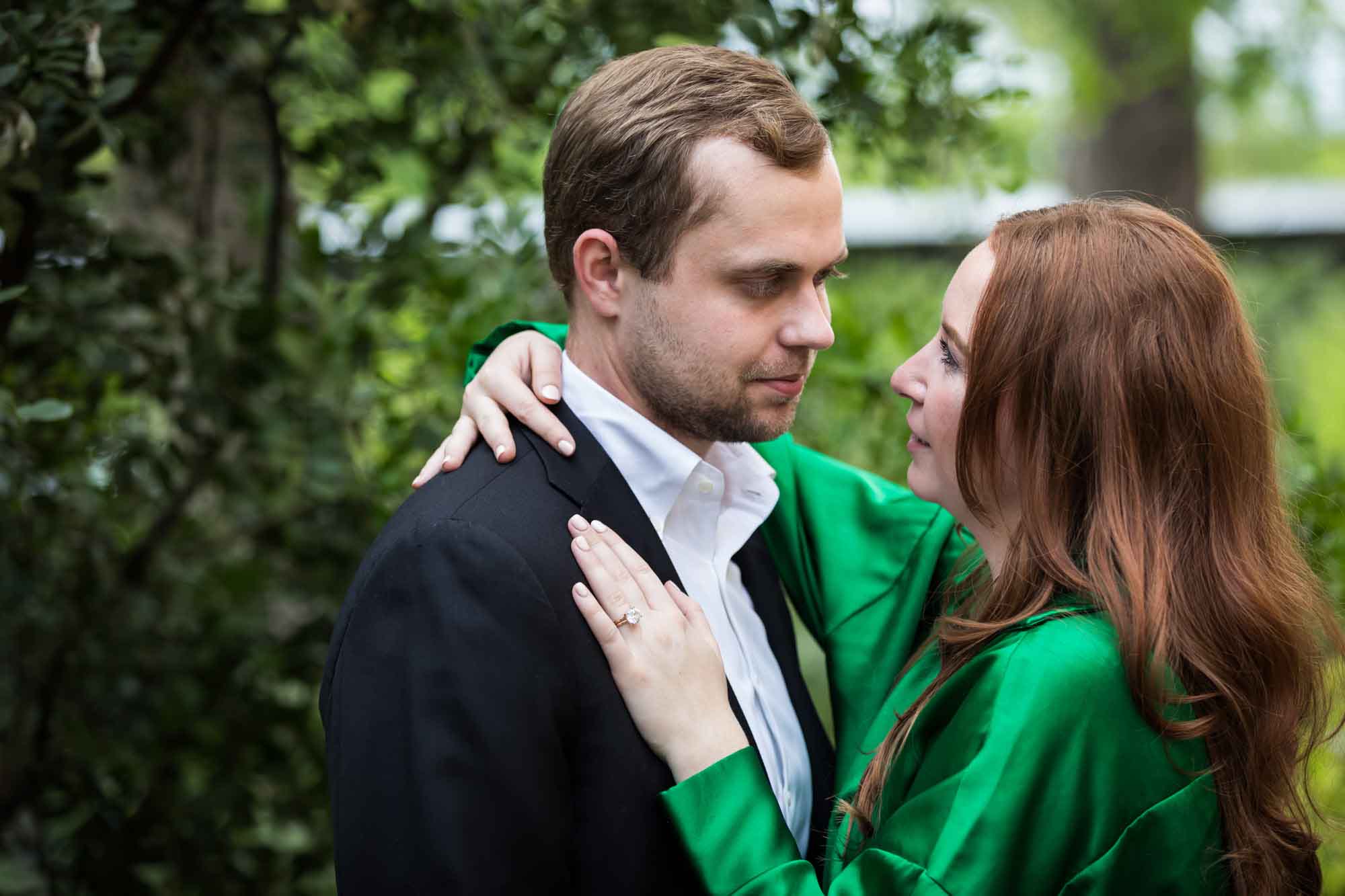 Man wearing black jacket and white shirt being hugged by woman wearing green blouse in front of green bushes during an Emma Hotel engagement photo shoot