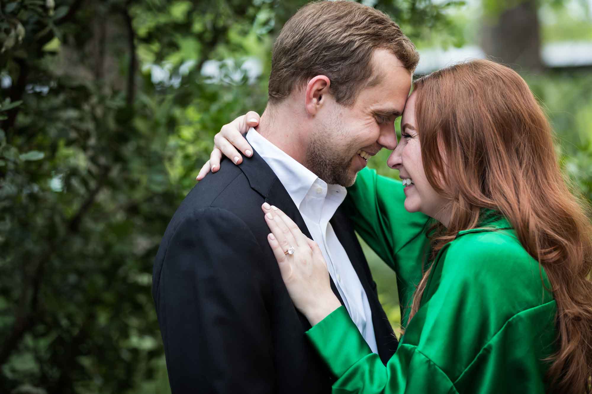 Man wearing black jacket and white shirt standing touching head to head with woman wearing green blouse in front of green bushes during an Emma Hotel engagement photo shoot