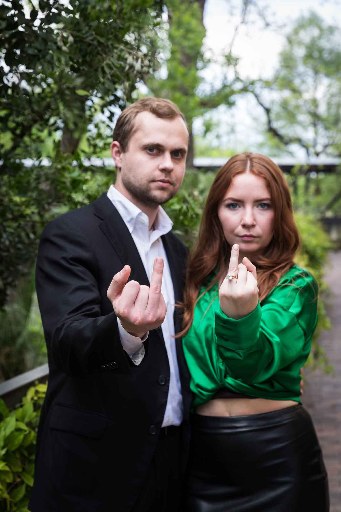 Man wearing black jacket and white shirt standing with woman wearing green blouse in front of green bushes with both people showing fingers to the camera and straight faces during an Emma Hotel engagement photo shoot