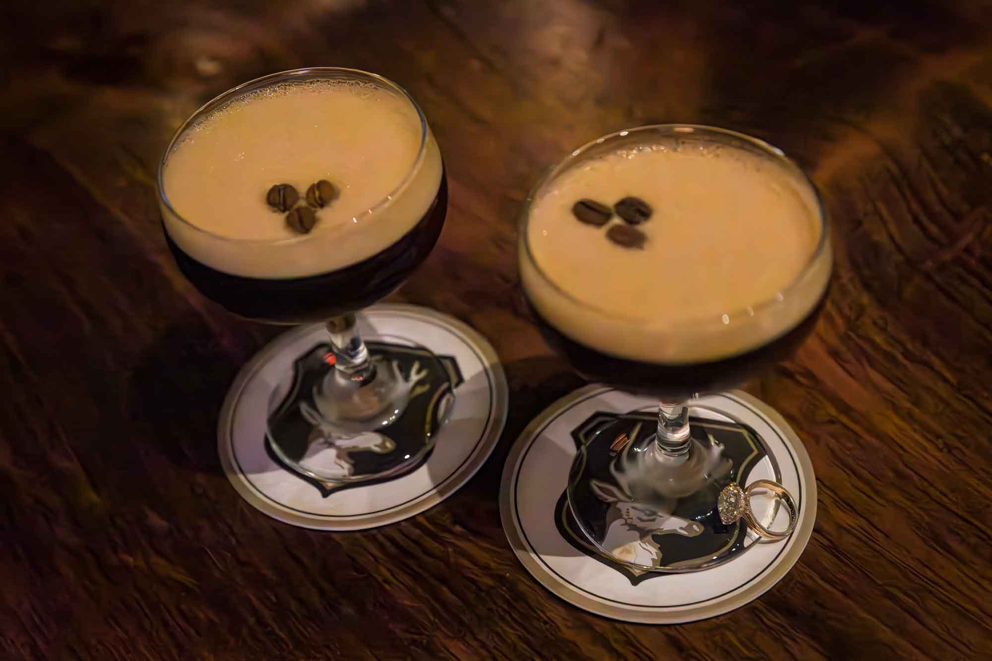 Close up of two martini glasses with espresso martinis on wooden table with an engagement ring at the base of one martini glass during an Emma Hotel engagement photo shoot