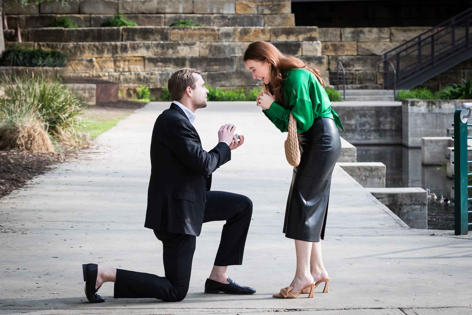 Man wearing black suit proposing while down on one knee in front of woman wearing green blouse on sidewalk in front of Emma Hotel beside Riverwalk during an Emma Hotel surprise proposal