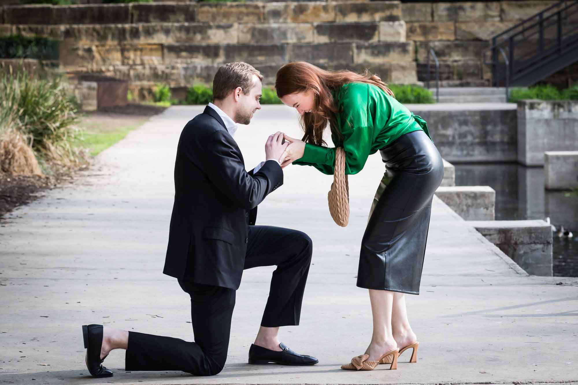 Man wearing black suit proposing while down on one knee in front of woman wearing green blouse on sidewalk in front of Emma Hotel beside Riverwalk during an Emma Hotel surprise proposal