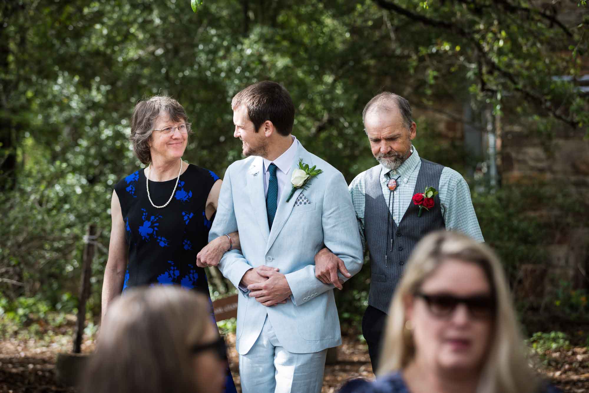 Groom walking arm in arm with both parents in front of guests during a Lady Bird Johnson Wildflower Center wedding