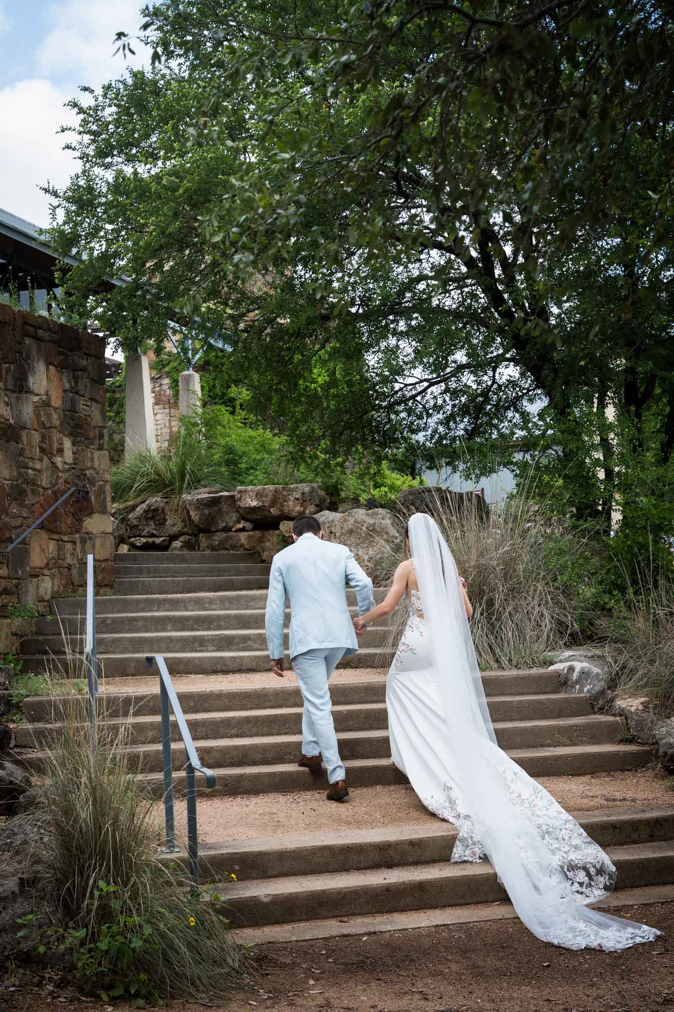 Bride and groom holding hands and walking up stairs with long veil trailing over stairs during a Lady Bird Johnson Wildflower Center wedding