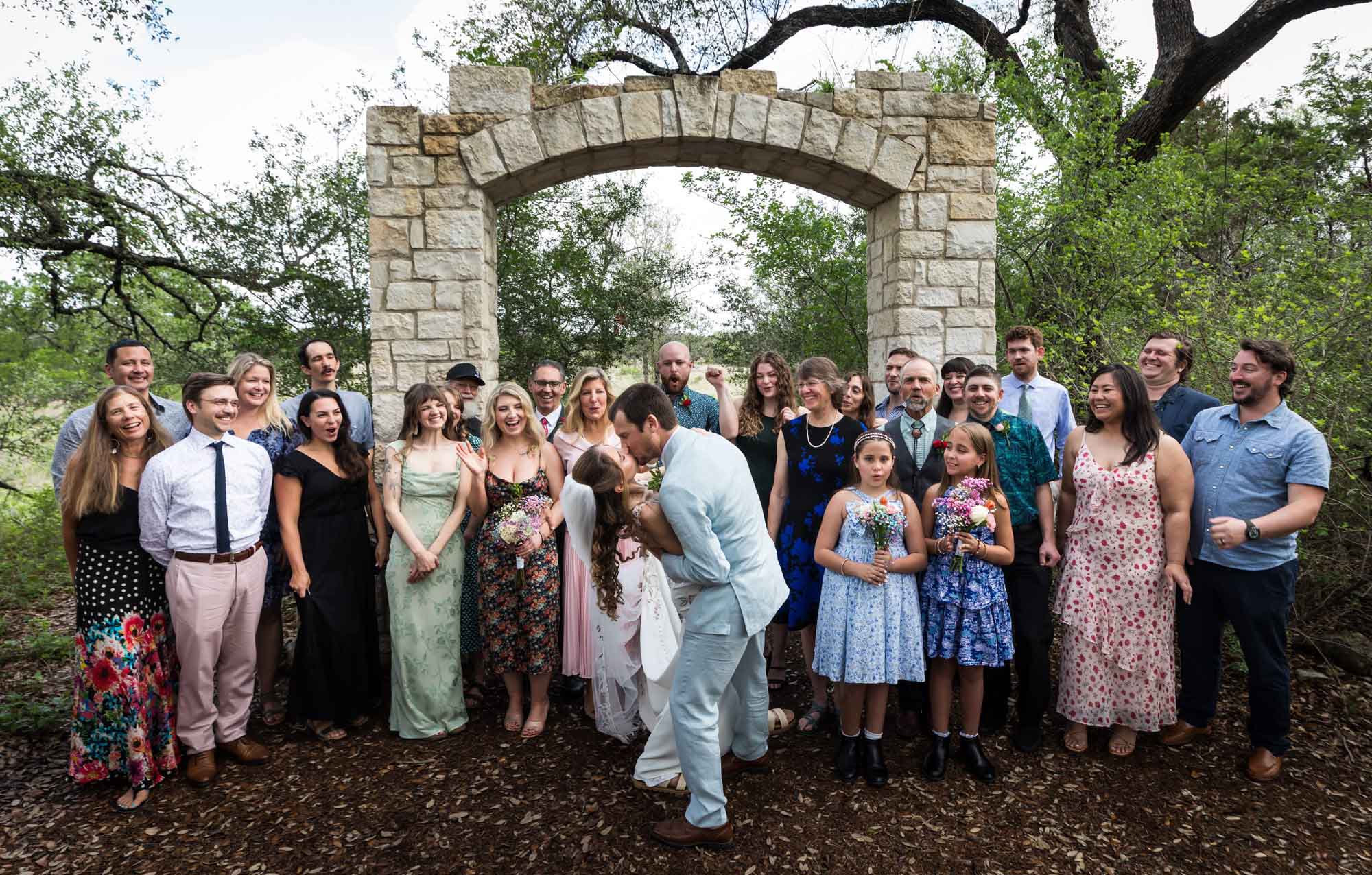 Large group photo of wedding guests cheering as bride and groom kiss in front of Cecille's Arch during a Lady Bird Johnson Wildflower Center wedding