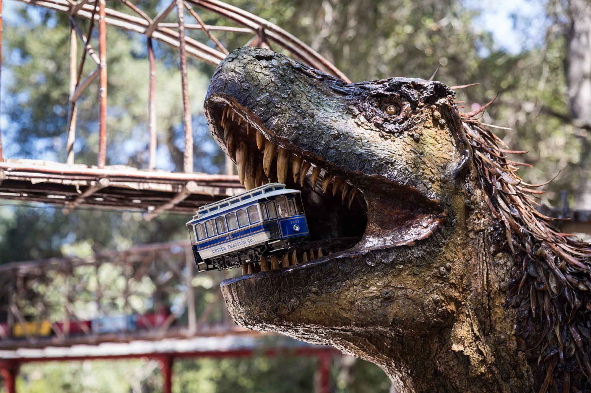 Wooden dinosaur sculpture holding a small train sculpture in its mouth in front of miniature train display for an article on the best photo shoot locations in Descanso Gardens