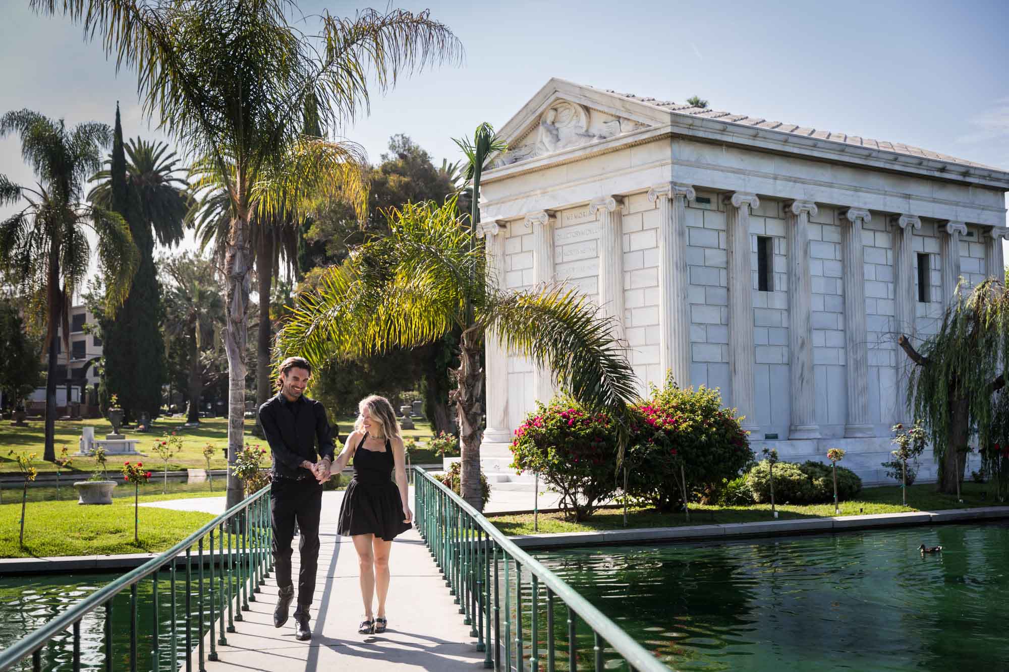 Hollywood Forever Cemetery engagement portrait of man and woman walking down pathway with Clark Mausoleum and lake in background