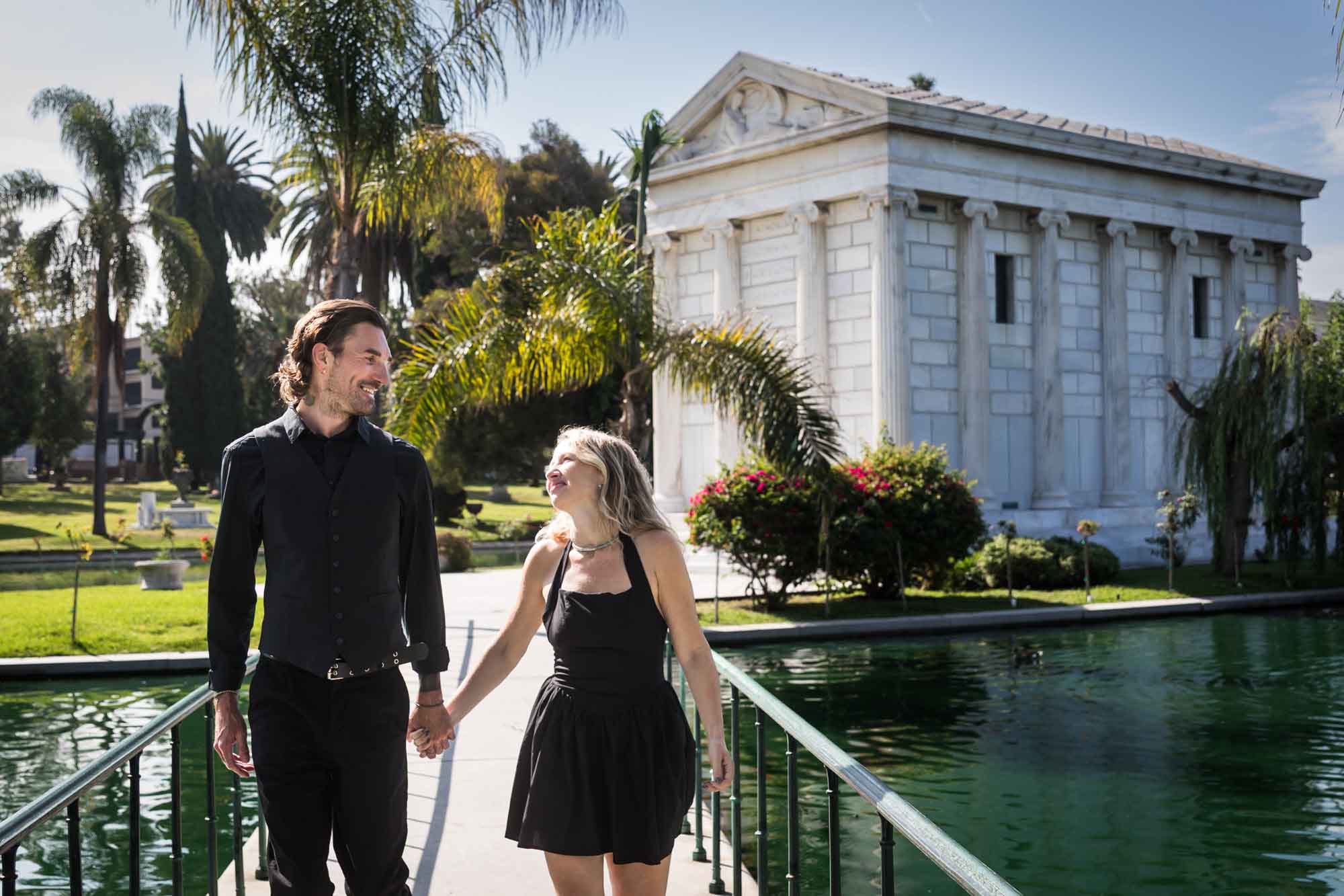 Hollywood Forever Cemetery engagement portrait of man and woman walking down pathway with Clark Mausoleum and lake in background