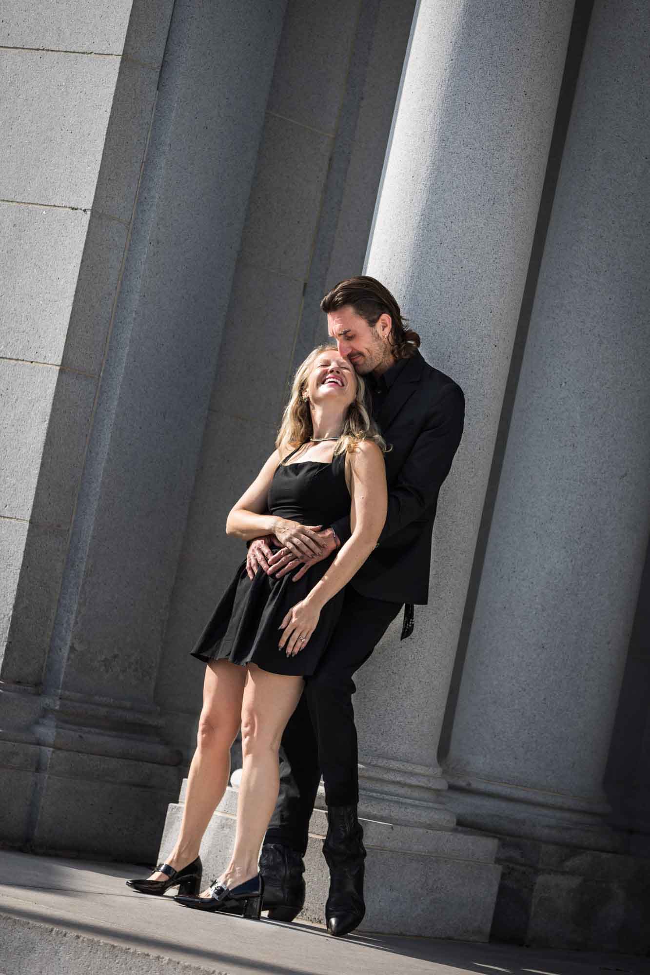 Man hugging woman from behind while standing next to stone pillar during a Hollywood Forever Cemetery engagement portrait session