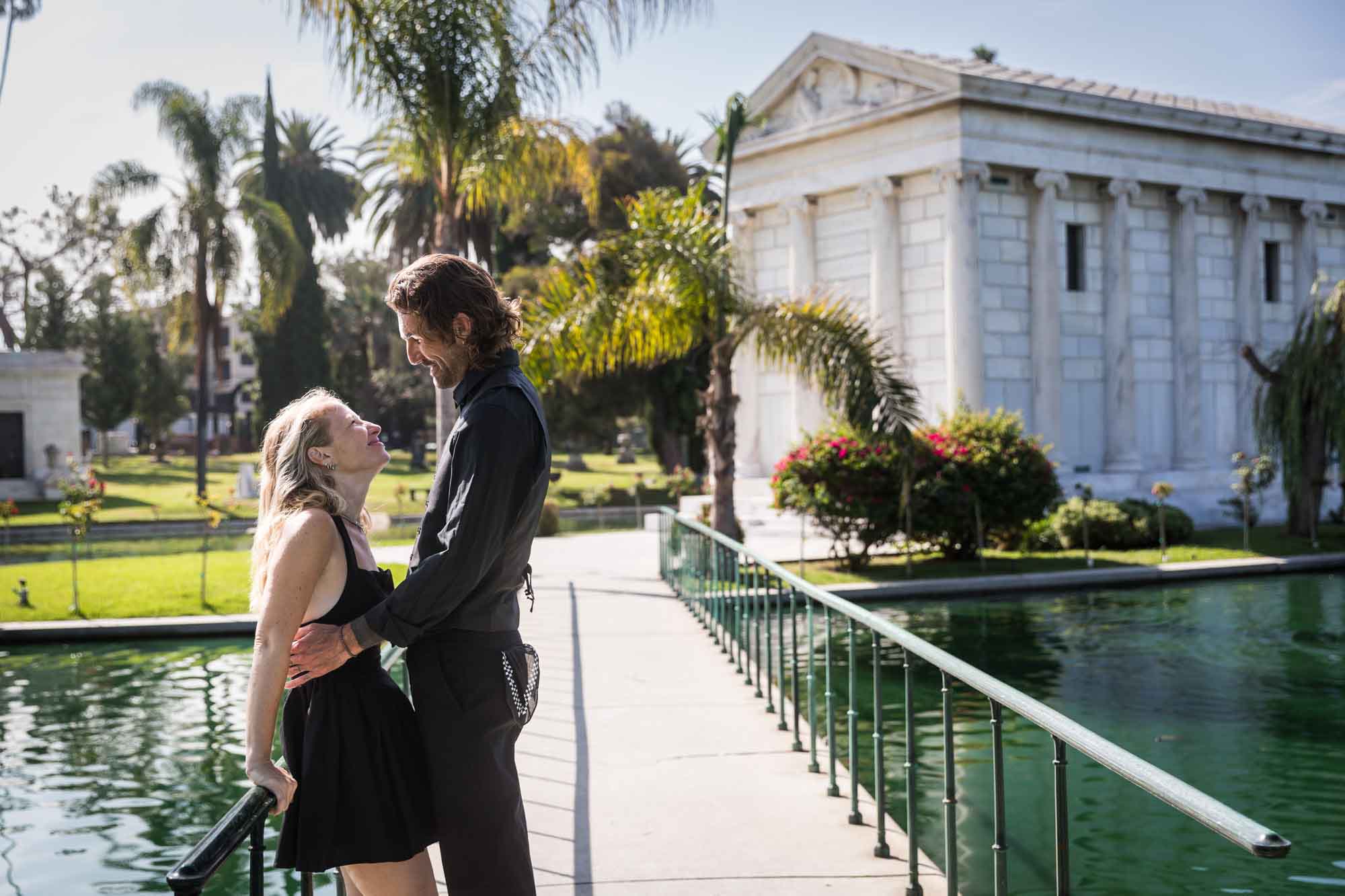 Hollywood Forever Cemetery engagement portrait of man and woman leaning against railing with lake and mausoleum in background