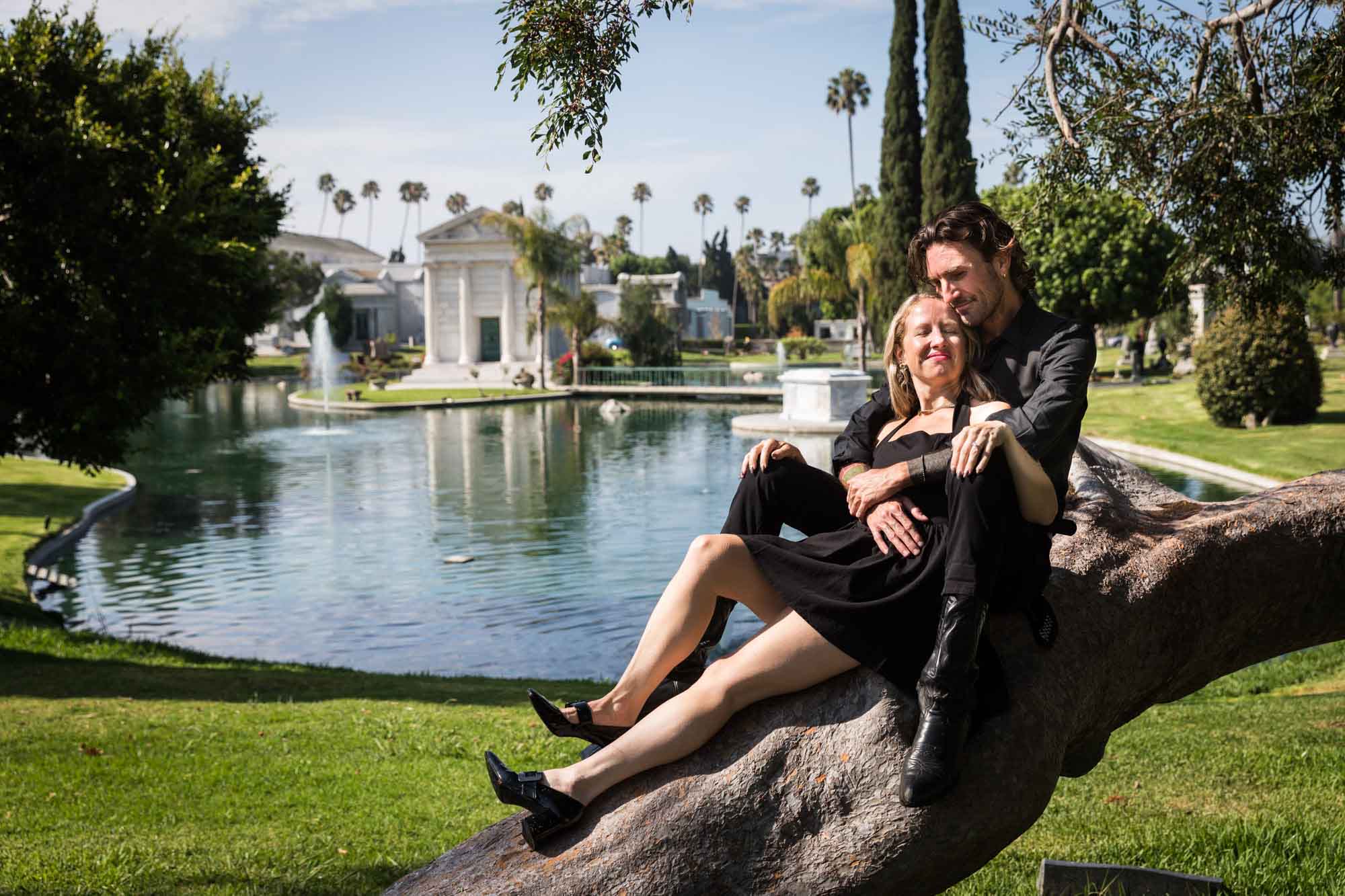 Hollywood Forever Cemetery engagement portrait of man and woman sitting on tree branch with Clark Mausoleum and lake in background