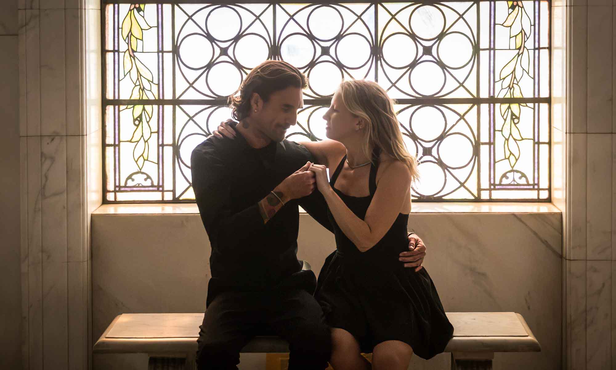 Hollywood Forever Cemetery engagement portrait of man looking at woman's hand while sitting in front of stained glass window in Cathedral Mausoleum