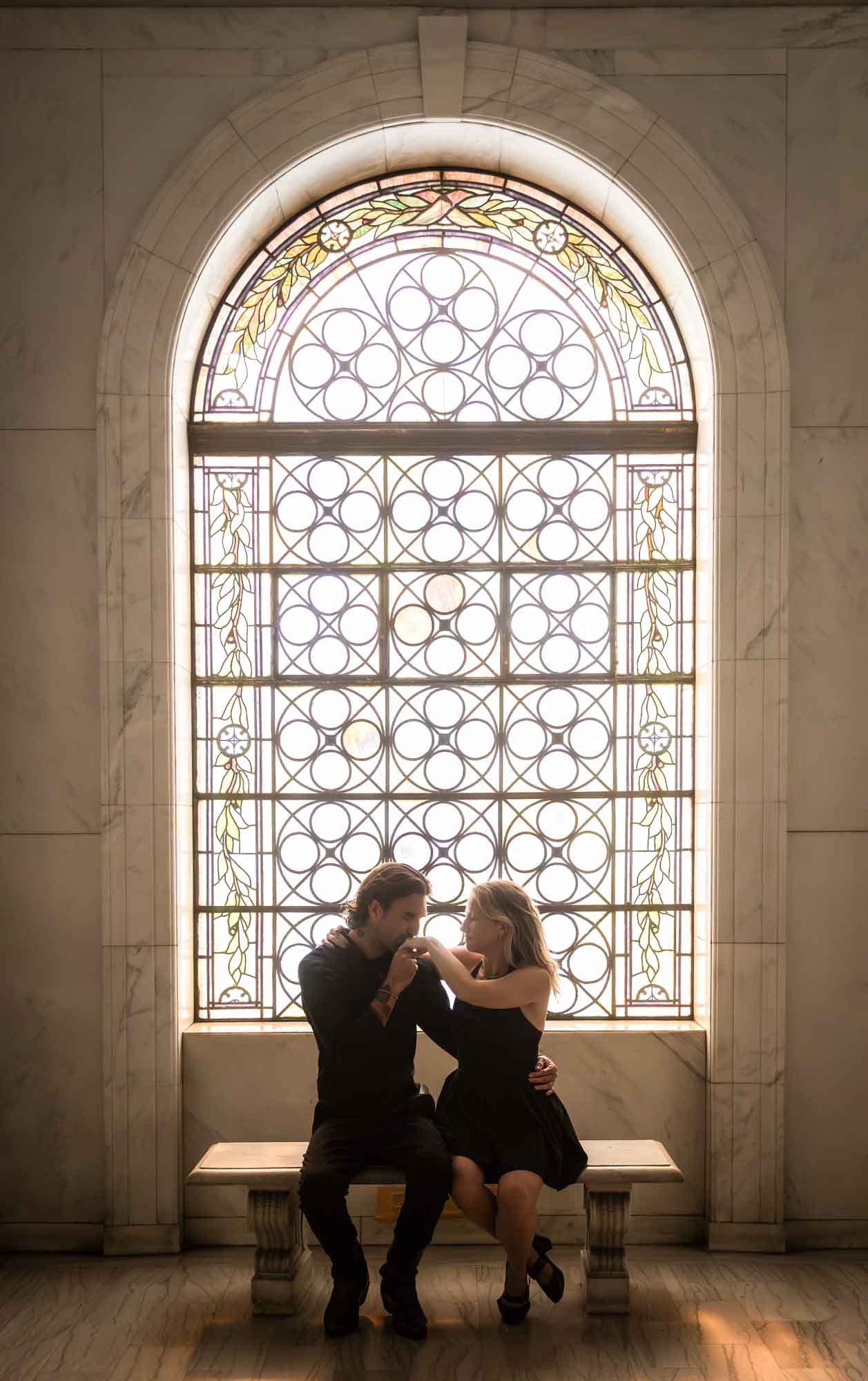 Hollywood Forever Cemetery engagement portrait of man kissing woman's hand while sitting in front of stained glass window in Cathedral Mausoleum