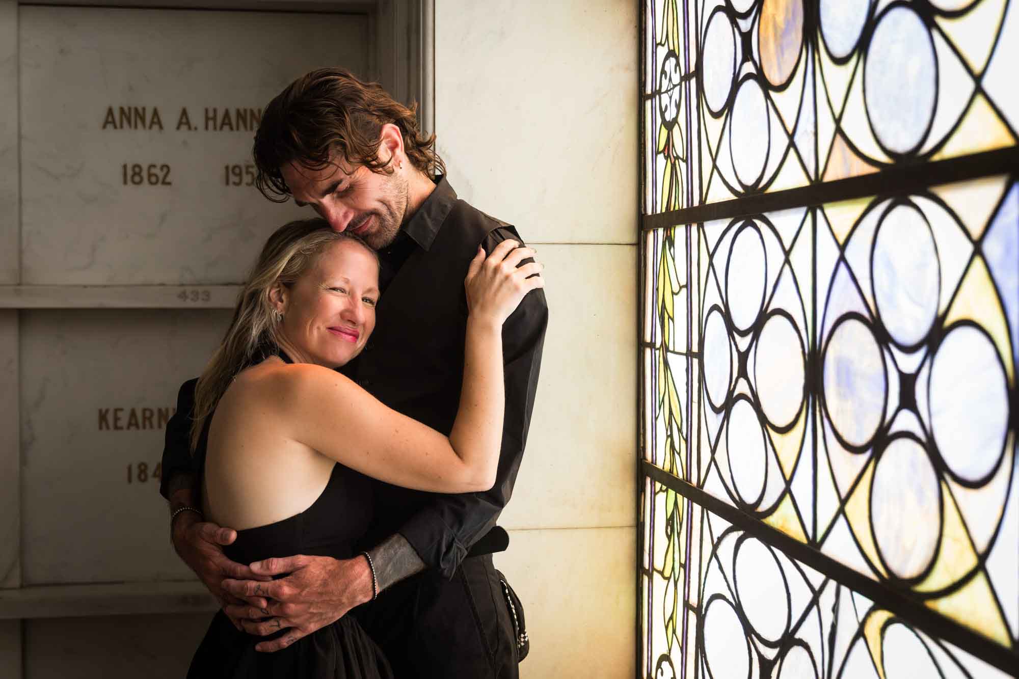 Hollywood Forever Cemetery engagement portrait of man and woman hugging in front of stained glass window in Cathedral Mausoleum
