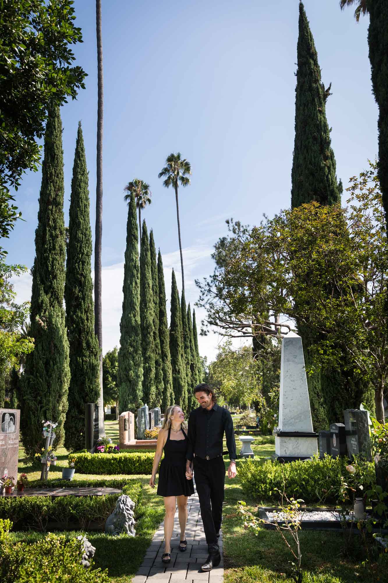 Man and woman wearing black outfits and holding hands while walking down Ancestral Forest walkway during a Hollywood Forever Cemetery engagement portrait session