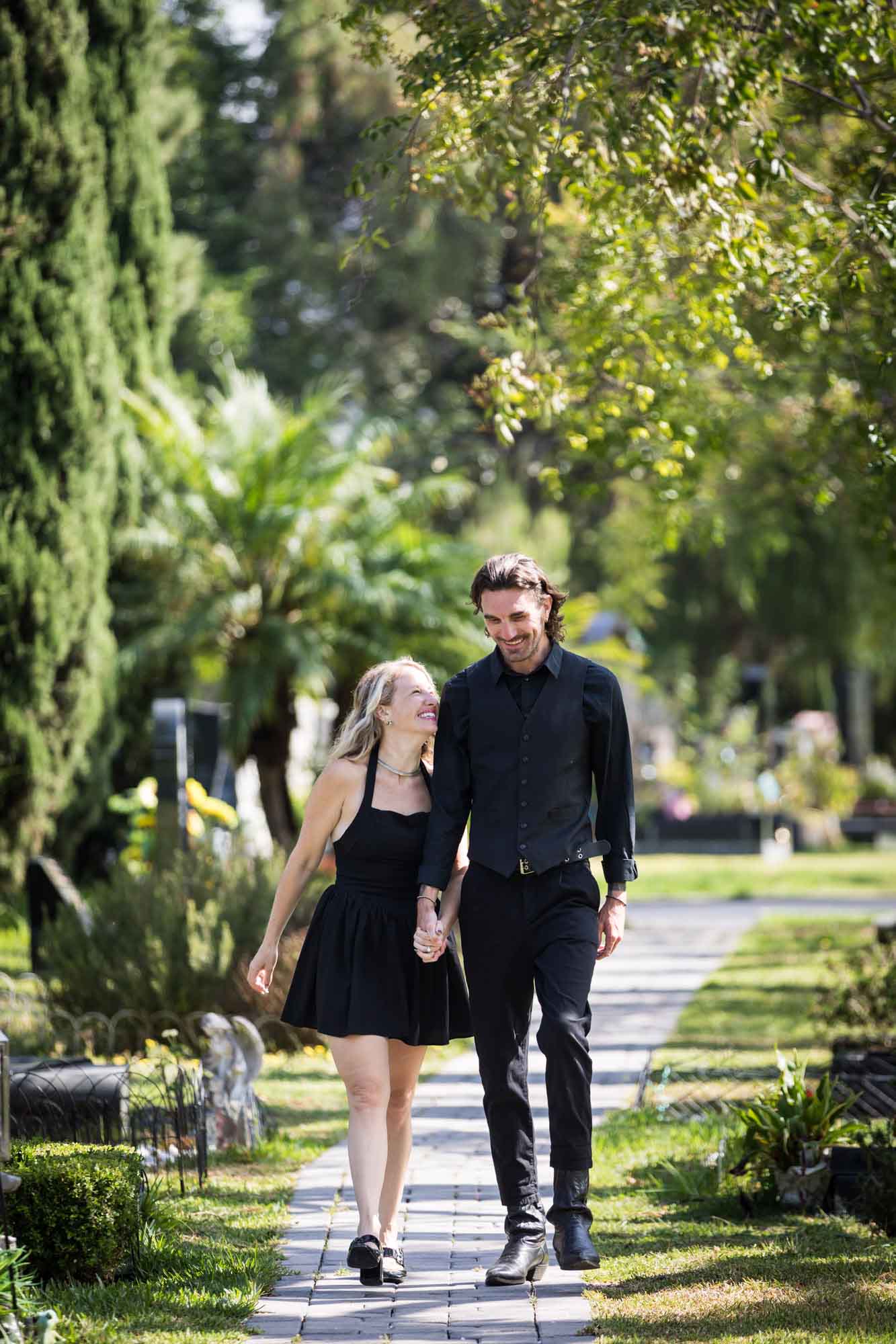Man and woman wearing black outfits and holding hands while walking down Ancestral Forest walkway during a Hollywood Forever Cemetery engagement portrait session