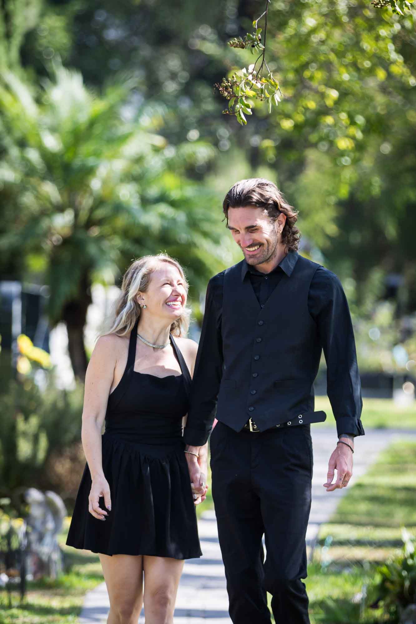 Man and woman wearing black outfits and holding hands while walking down Ancestral Forest walkway during a Hollywood Forever Cemetery engagement portrait session