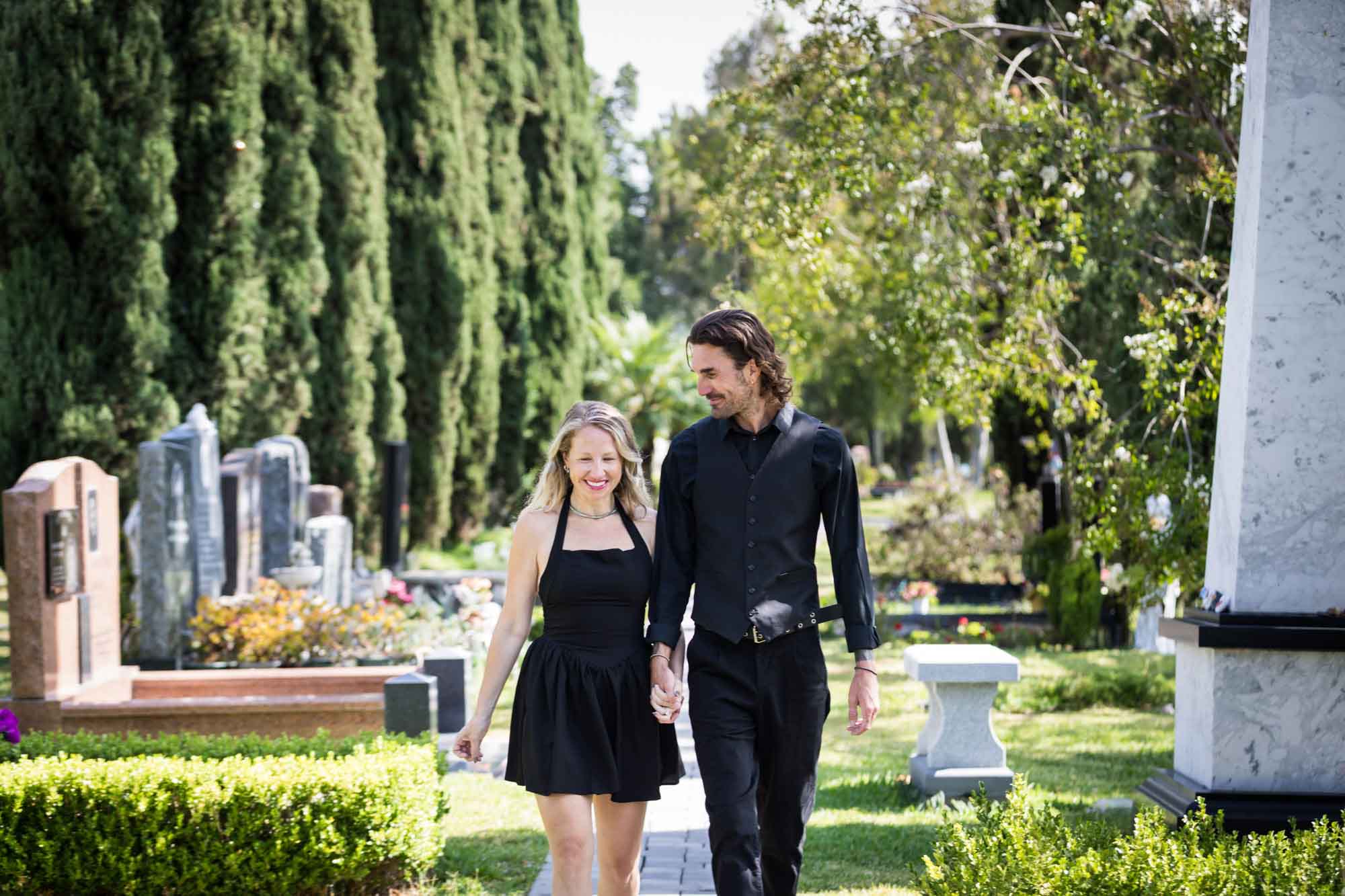 Man and woman wearing black outfits and holding hands while walking down Ancestral Forest walkway during a Hollywood Forever Cemetery engagement portrait session